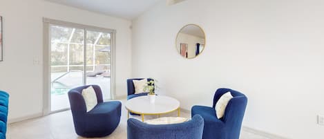 A modern living room with a round white table, four blue armchairs, a round mirror, and a glass door leading to a balcony.