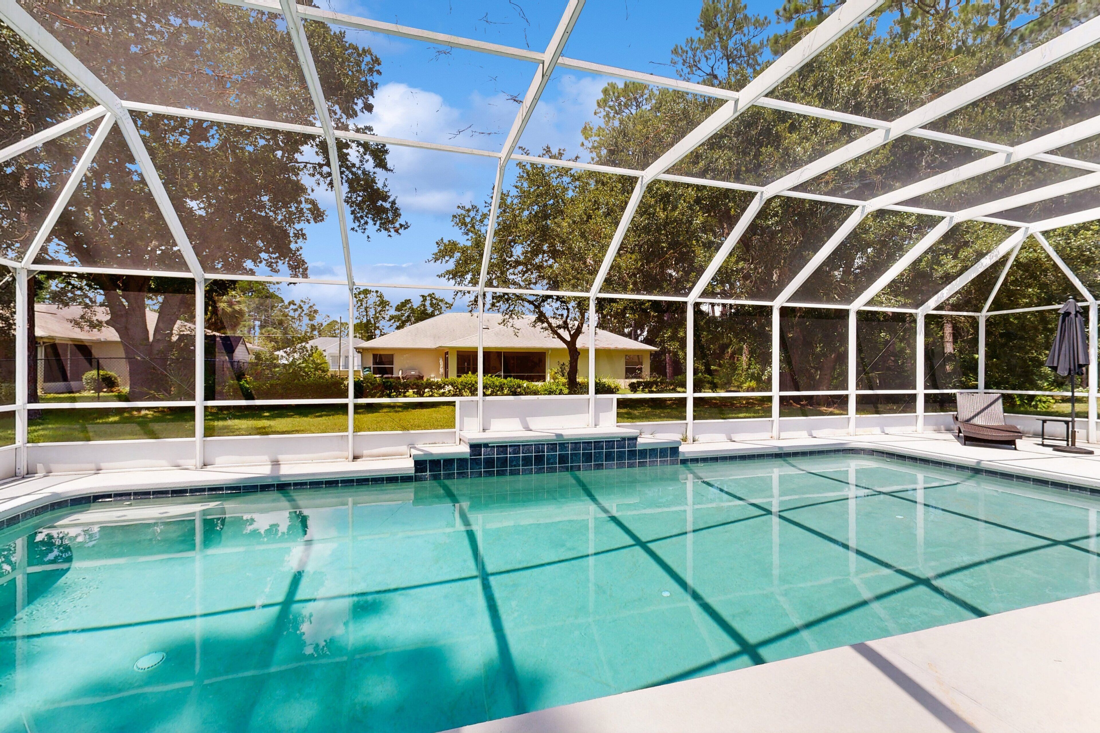 A clear blue pool with a white roofed enclosure, surrounded by trees and a house in the background.