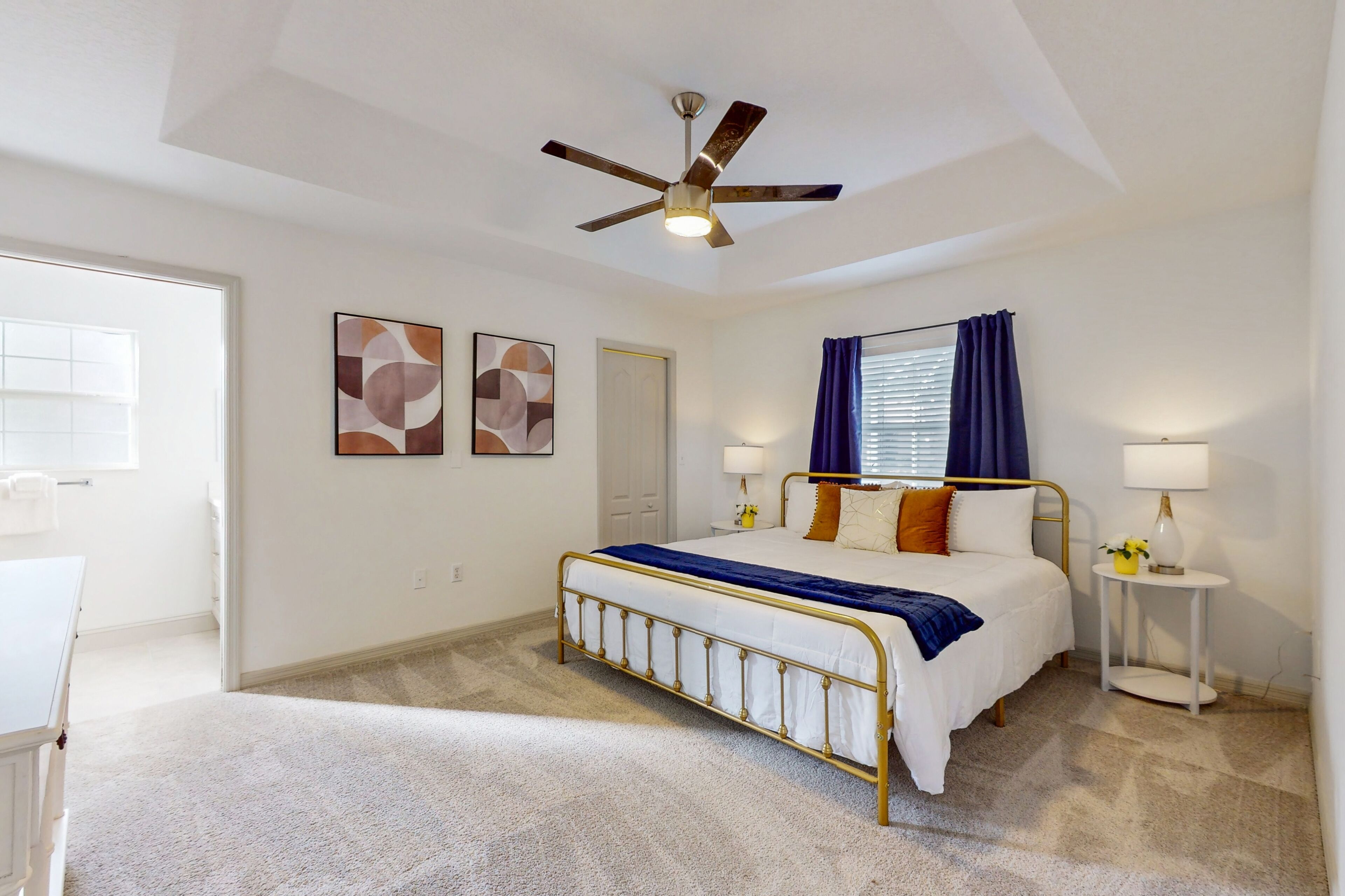 A bedroom with a bed, ceiling fan, artwork, bedside tables, and a window with curtains.