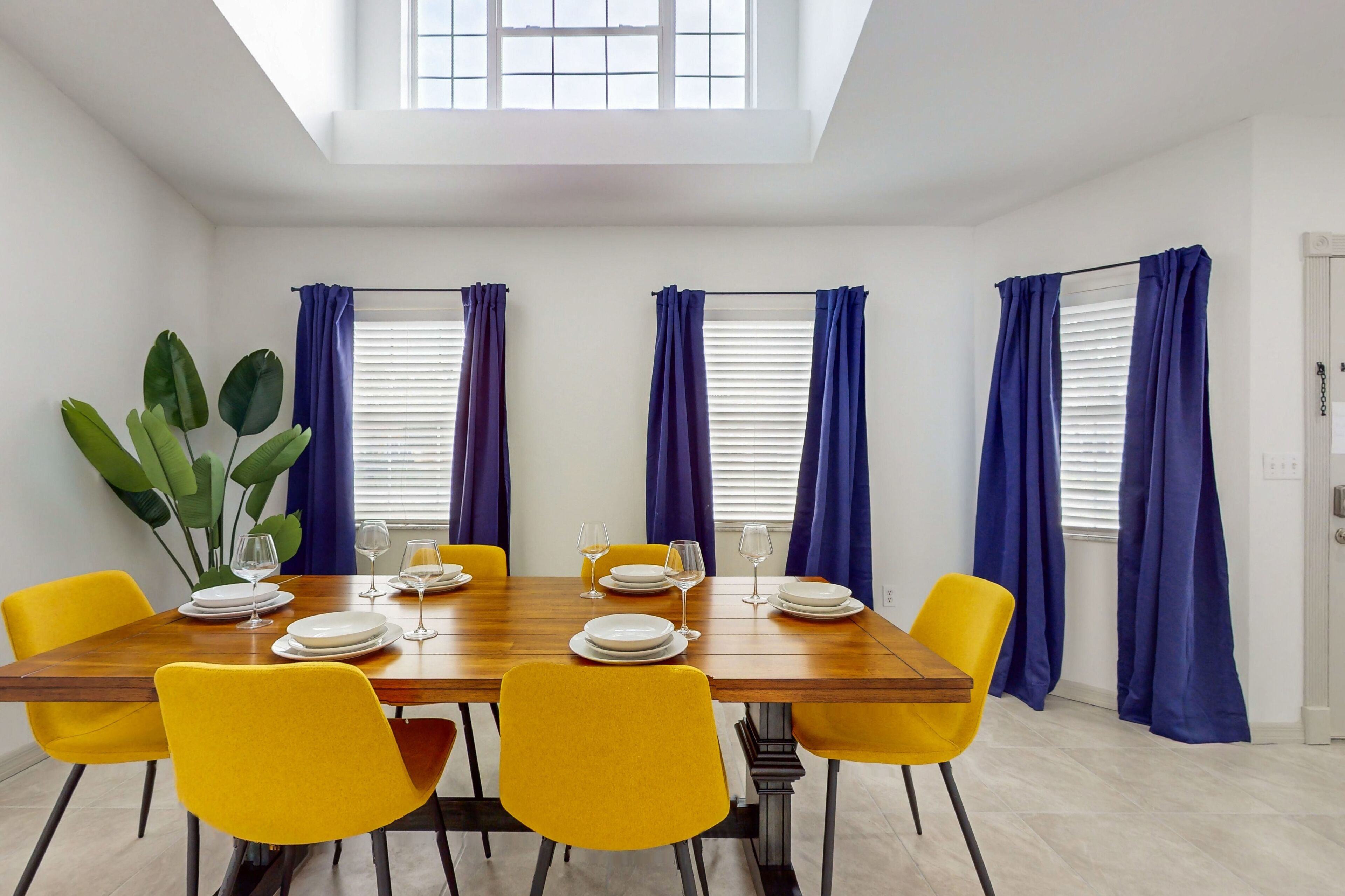 Comedor con mesa de madera dispuesta para seis comensales, sillas amarillas y cortinas azules.
