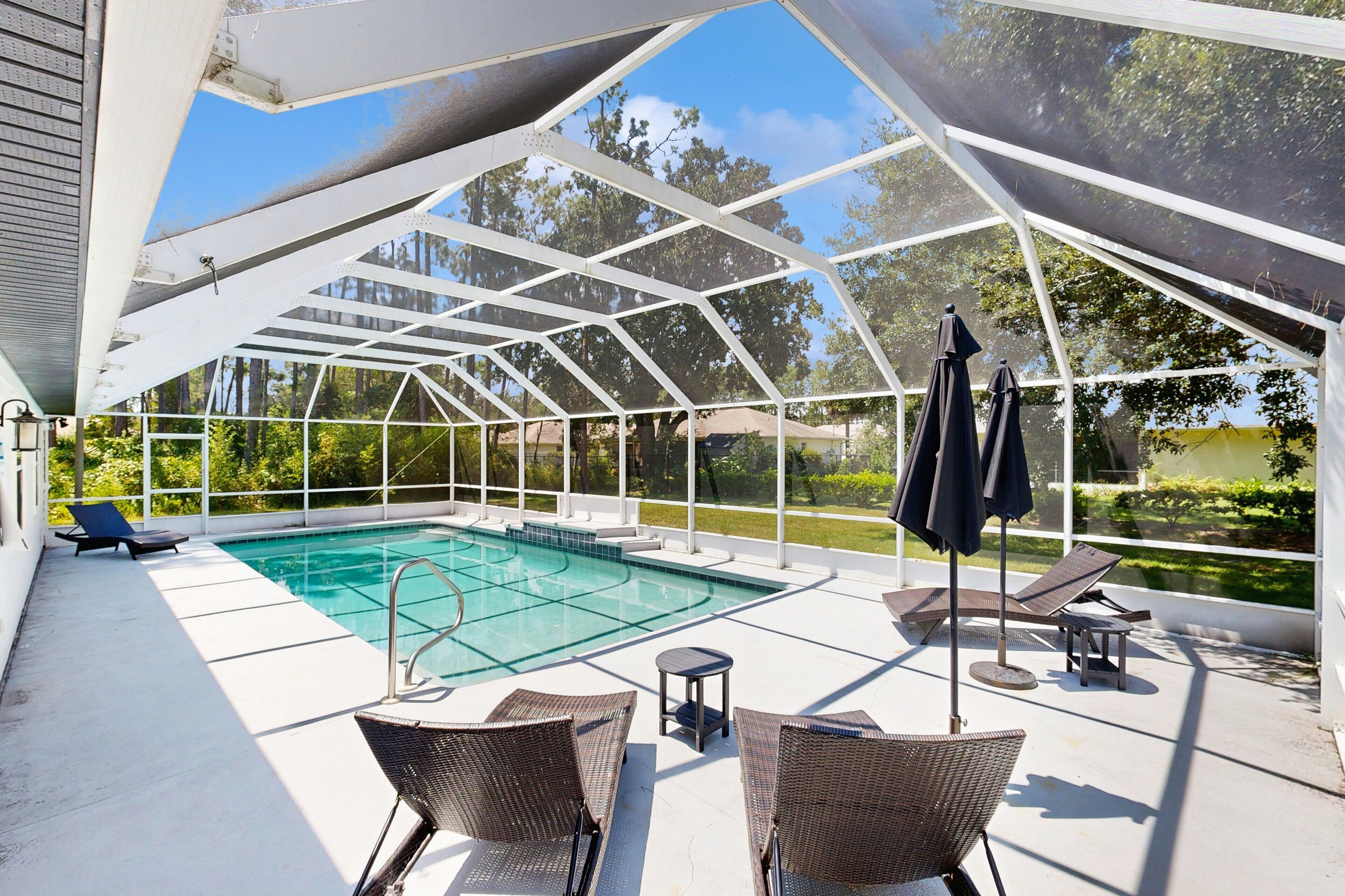 A covered outdoor swimming pool with lounge chairs and umbrellas, surrounded by a white metal frame and glass panels.