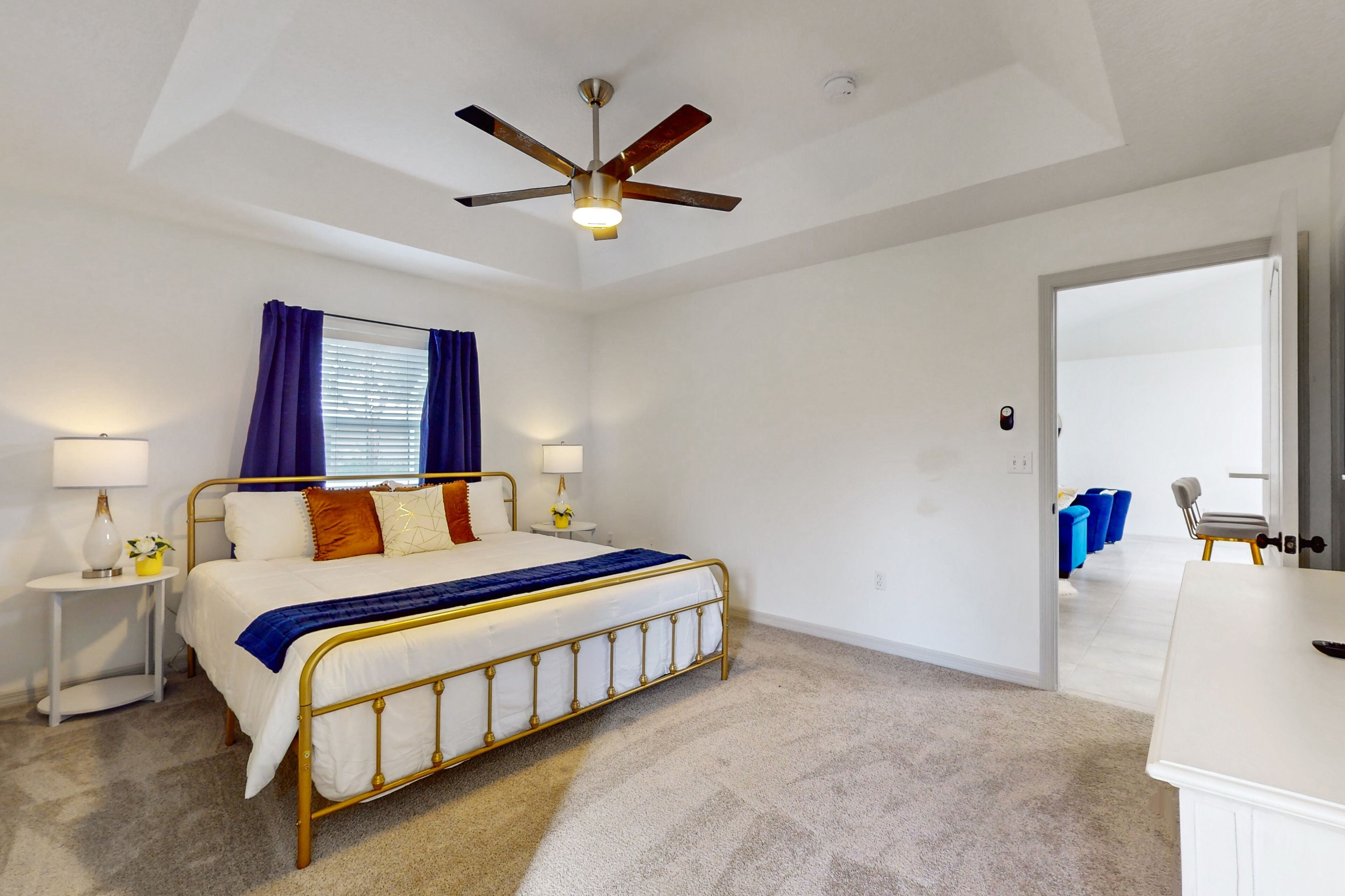 A bedroom with a bed, a ceiling fan, a nightstand with a lamp, and a view into another room with blue chairs.