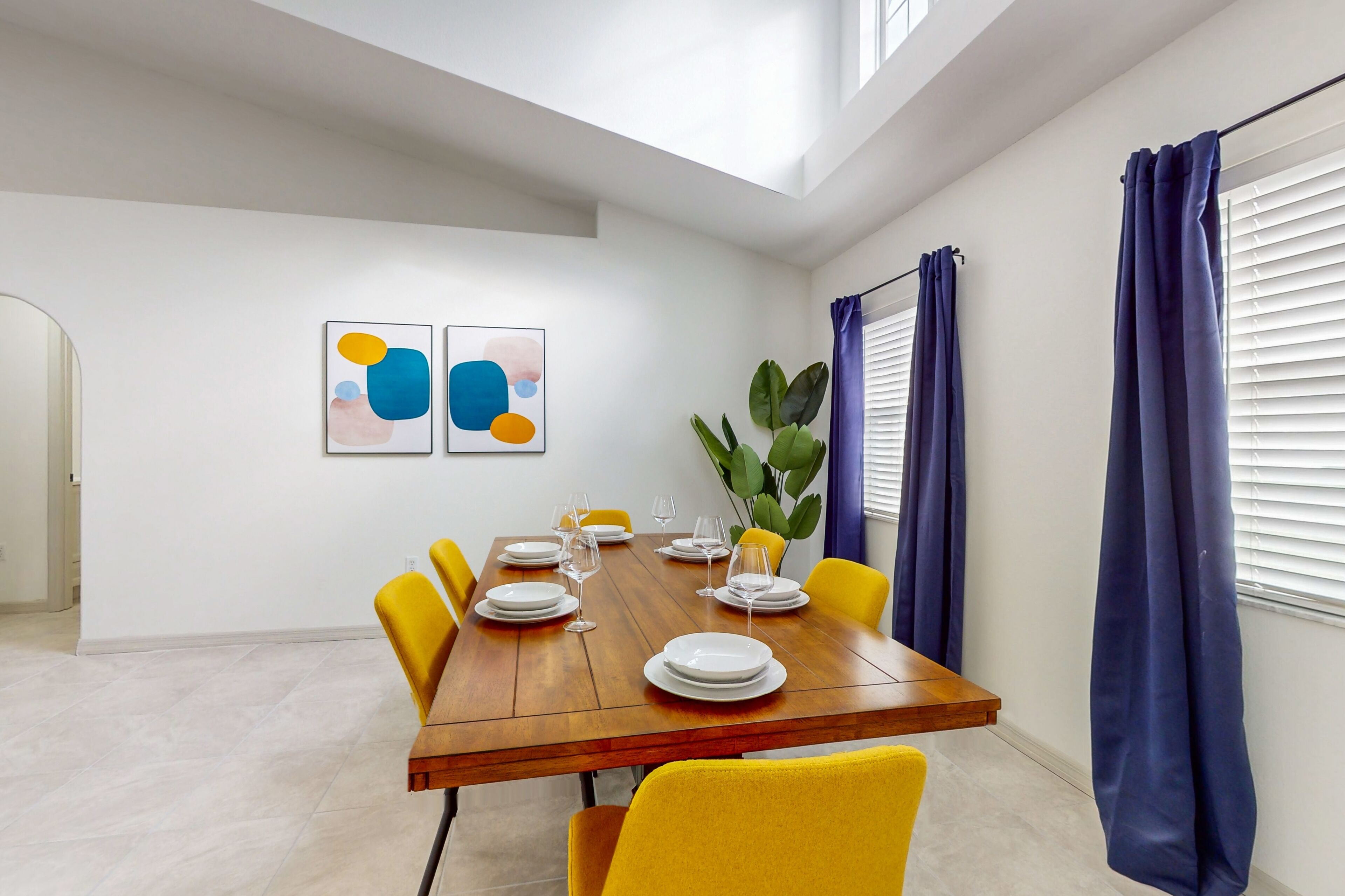 Un comedor con una mesa de madera dispuesta para seis comensales, sillas amarillas y una planta en maceta.