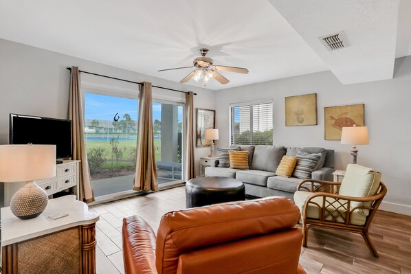 A spacious living room with a large window, a ceiling fan, a flat-screen TV, a sofa, a chair, and a coffee table. - Ground-floor Condo Next to the Tennis Courts With 2 Pools, Sauna, Beach Access (St. Augustine)