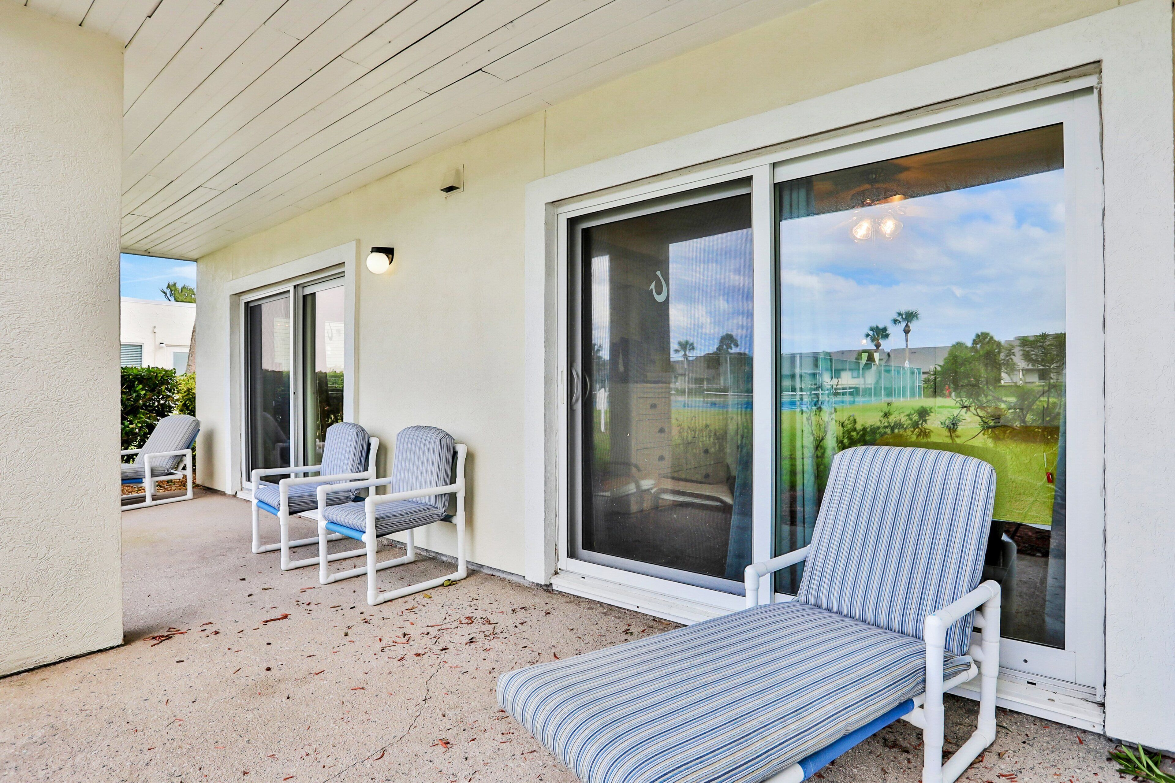 A covered outdoor area with two chairs and a cushioned bench, a sliding glass door, and a view of a green landscape.