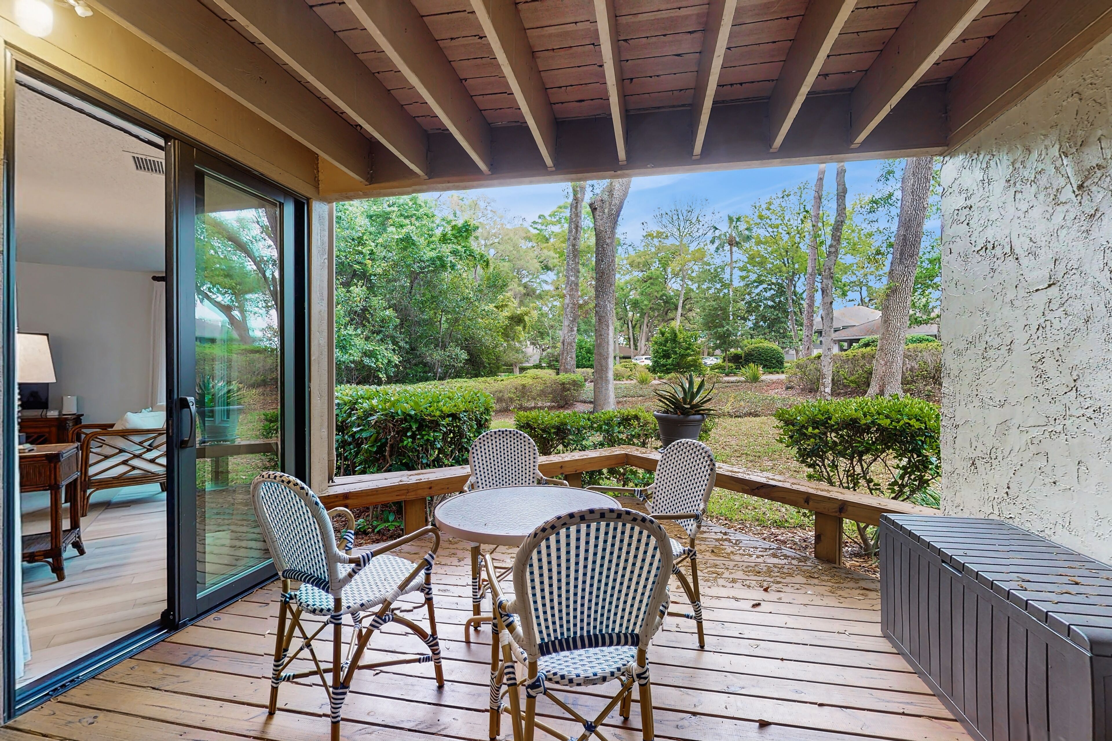 An outdoor patio with a wooden table and wicker chairs, a glass door leading to a room, and a view of a garden with trees and shrubs.