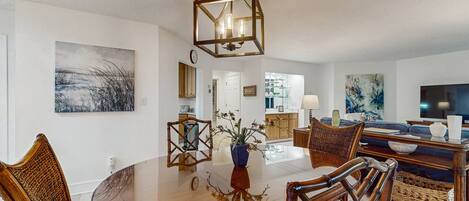 A dining area with a glass-top table, wooden chairs, a hanging light fixture, and artwork on the walls.