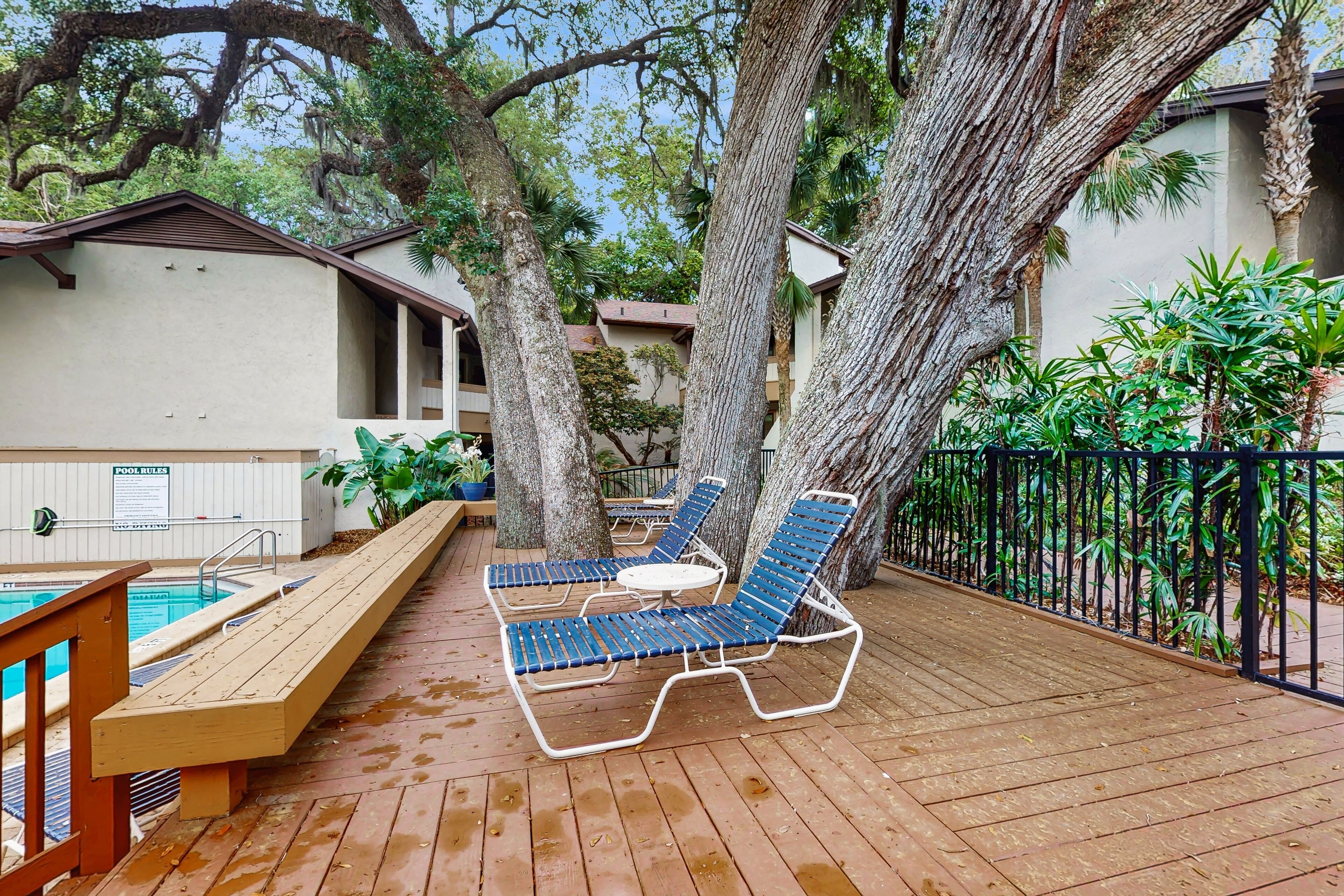 A wooden deck with two blue lounge chairs and a small table, a pool, and a sign reading "POOL RULES".