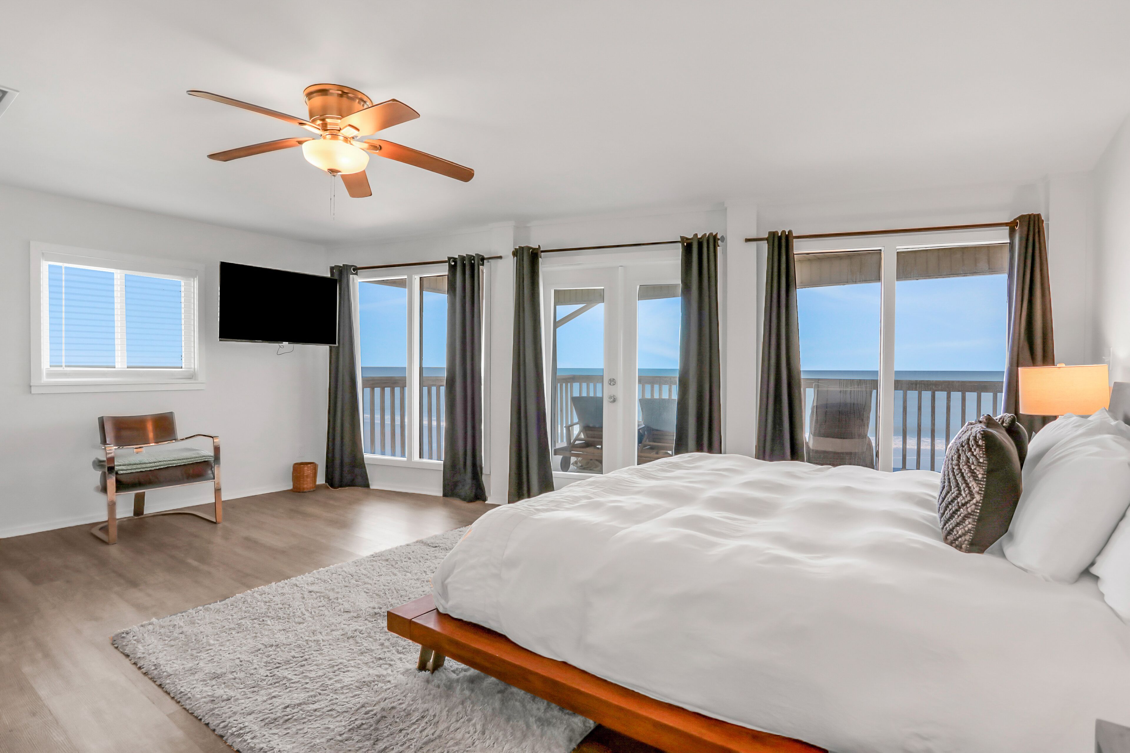 A bedroom with a large bed, a ceiling fan, a TV, and a view of the ocean through the windows.