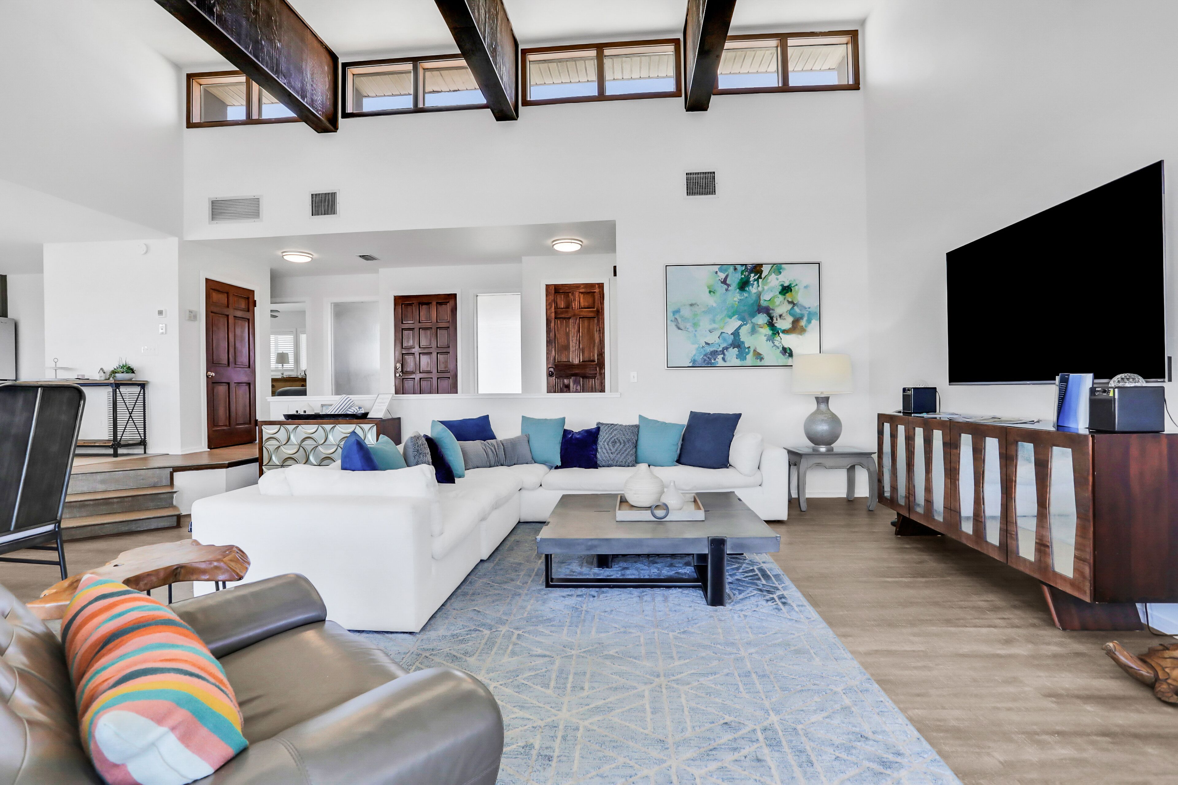A spacious living room with a white sectional sofa, a coffee table, and a TV stand. There are wooden doors and a staircase leading to another room.