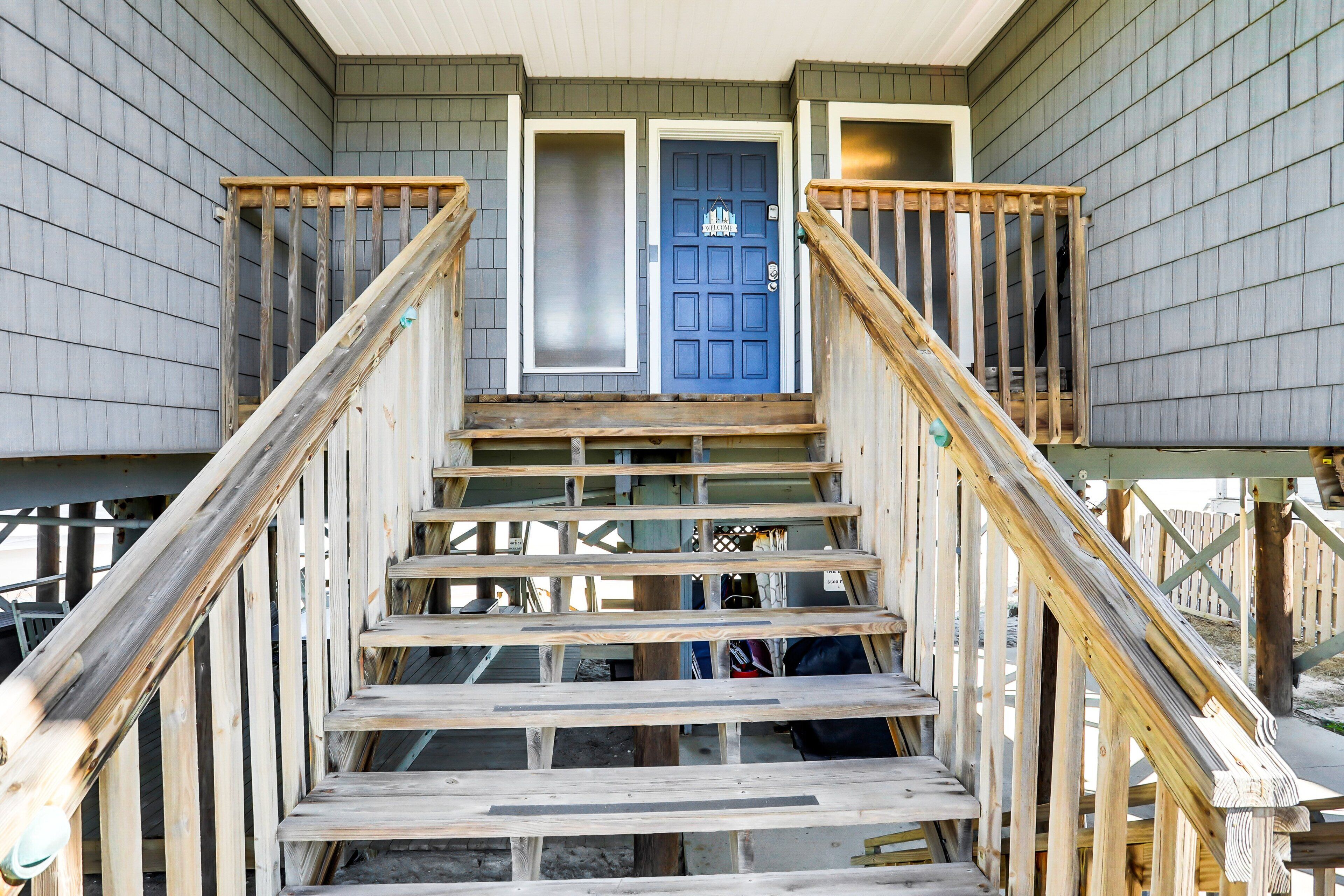 A wooden staircase with a blue door at the top, leading to a covered area with a wooden railing.