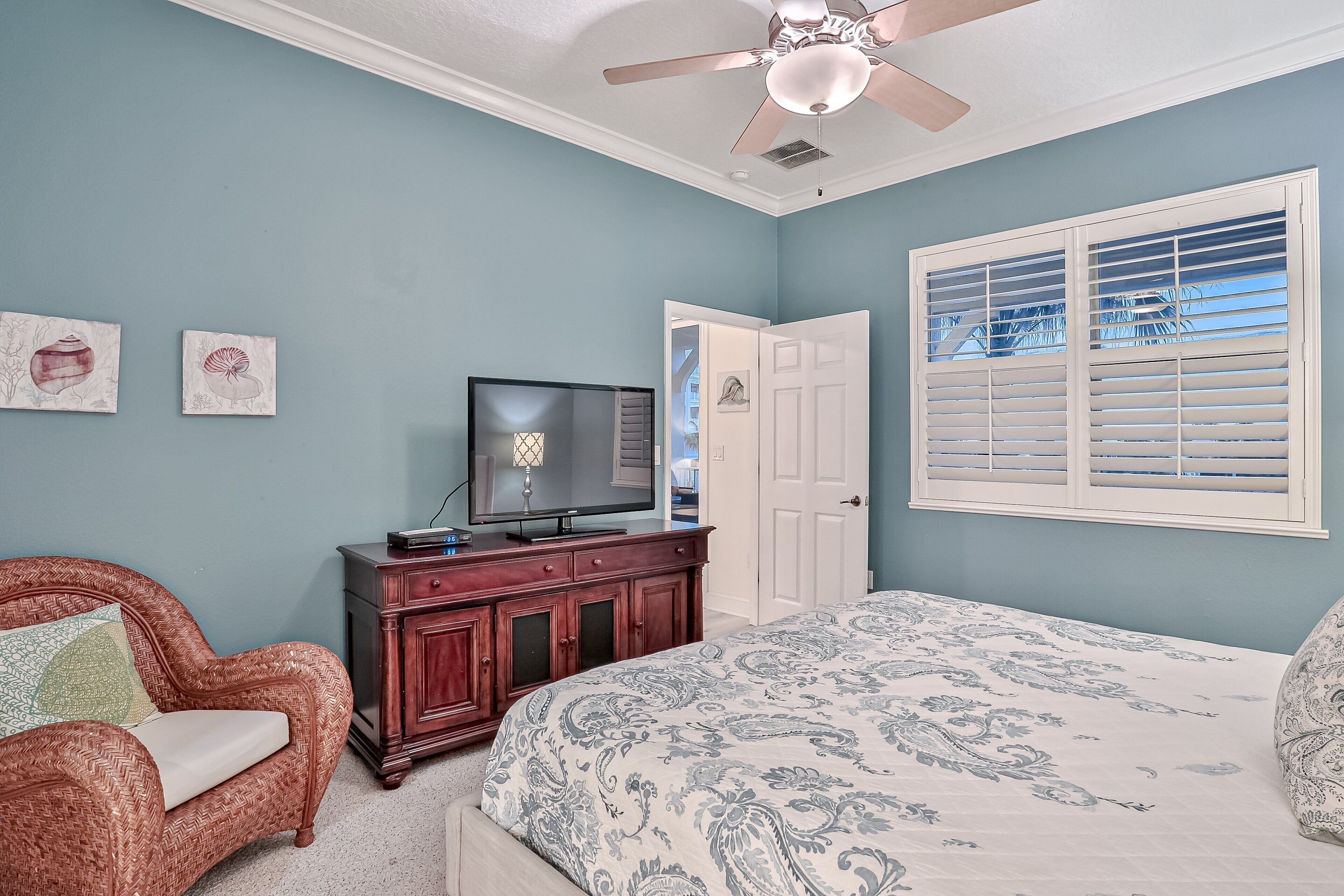 A bedroom with a bed, a television on a wooden cabinet, a ceiling fan, and a window with blinds.