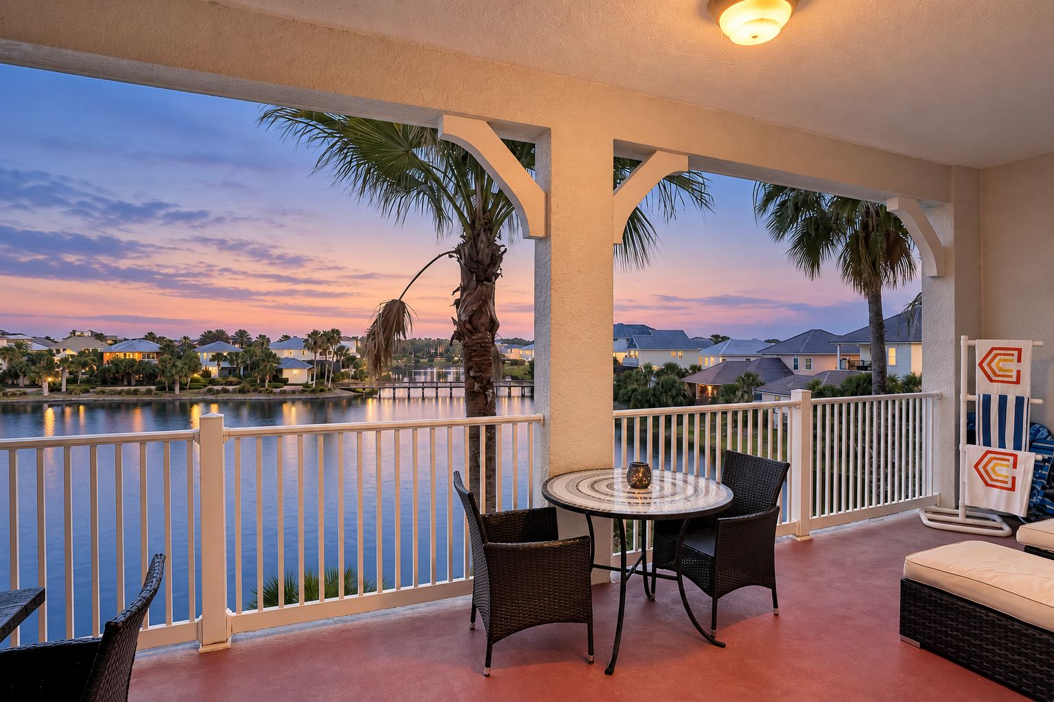 A balcony with a view of a lake and houses, furnished with wicker chairs and a table, and a railing with decorative towel holders.
