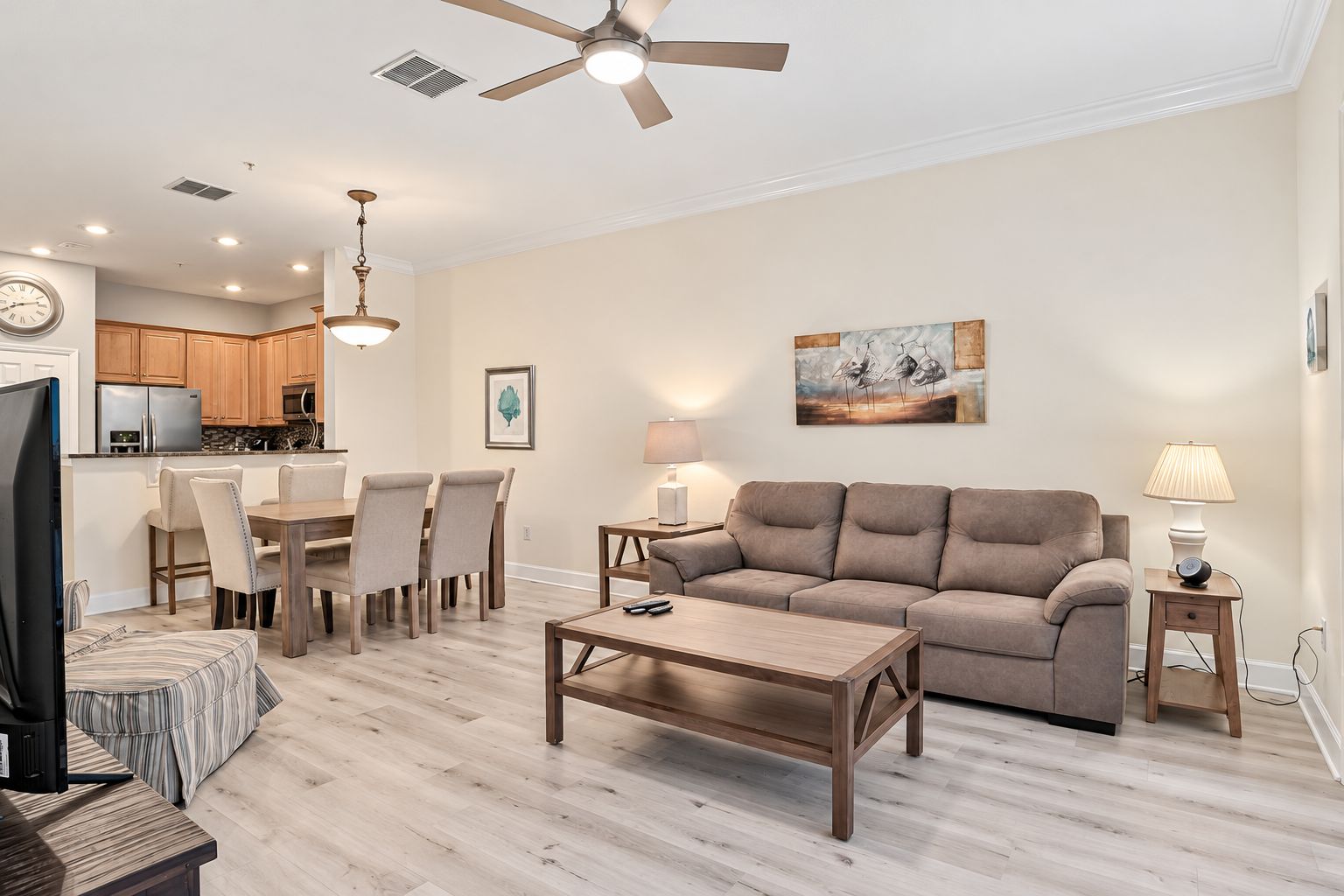A living room with a sofa, coffee table, and TV. There is a dining area with chairs and a table. The room has wooden flooring and a ceiling fan.
