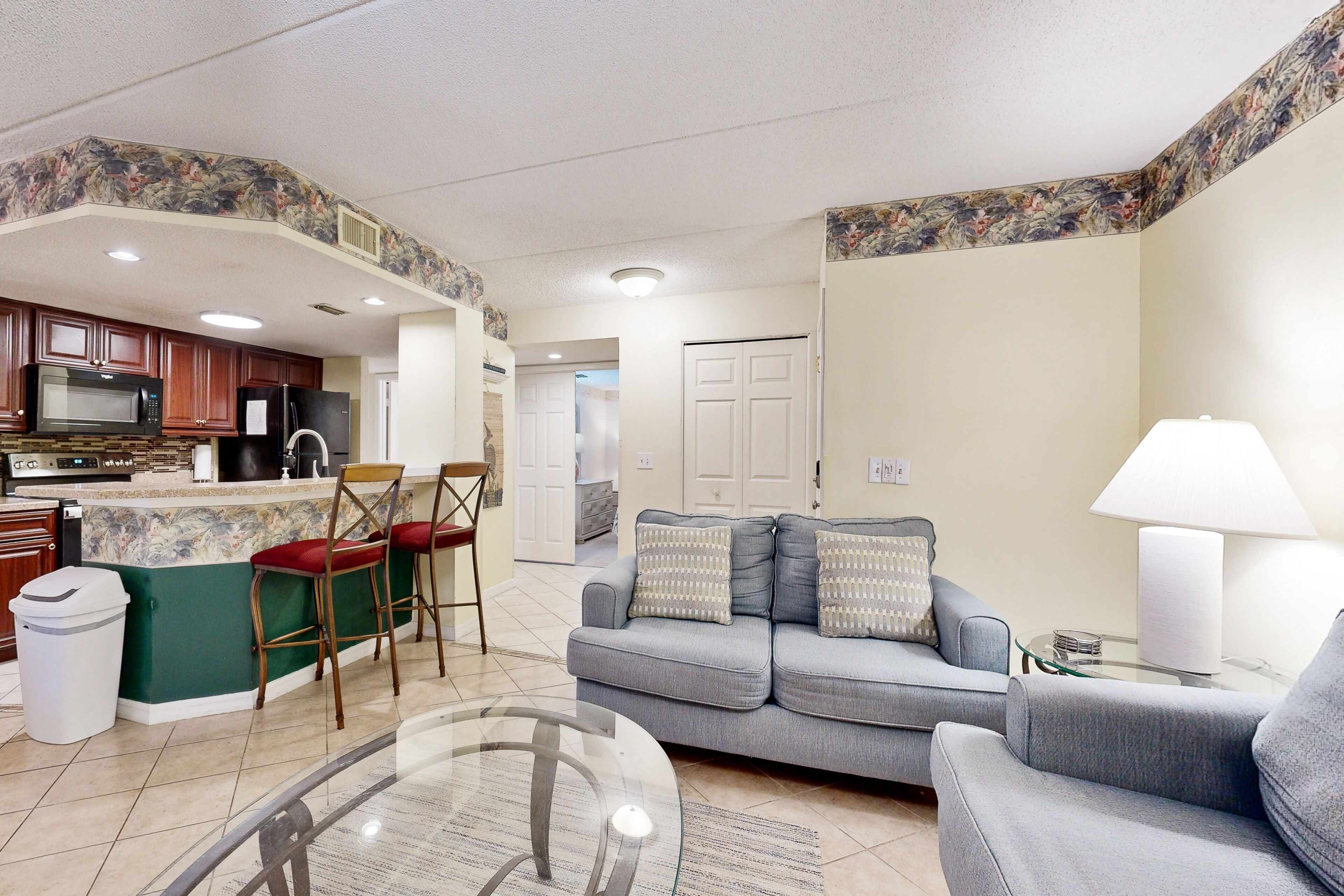 A modern kitchen with a microwave, refrigerator, and bar stools. There is a gray sofa with patterned cushions in the living area.