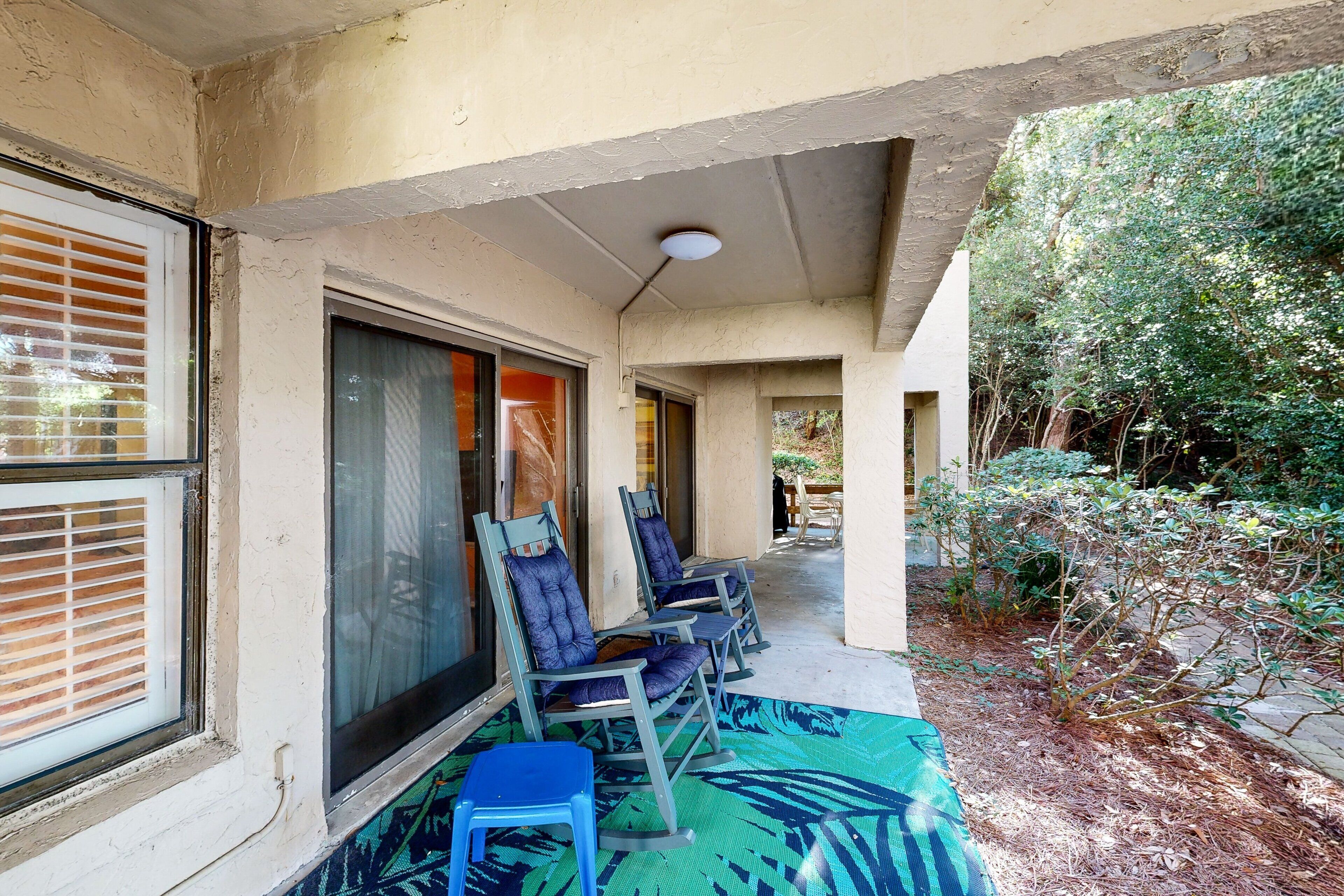 Un espace extérieur aménagé, abrité et doté de chaises et d’une table, une fenêtre avec des stores et une vue sur un jardin avec des plantes et un sentier.