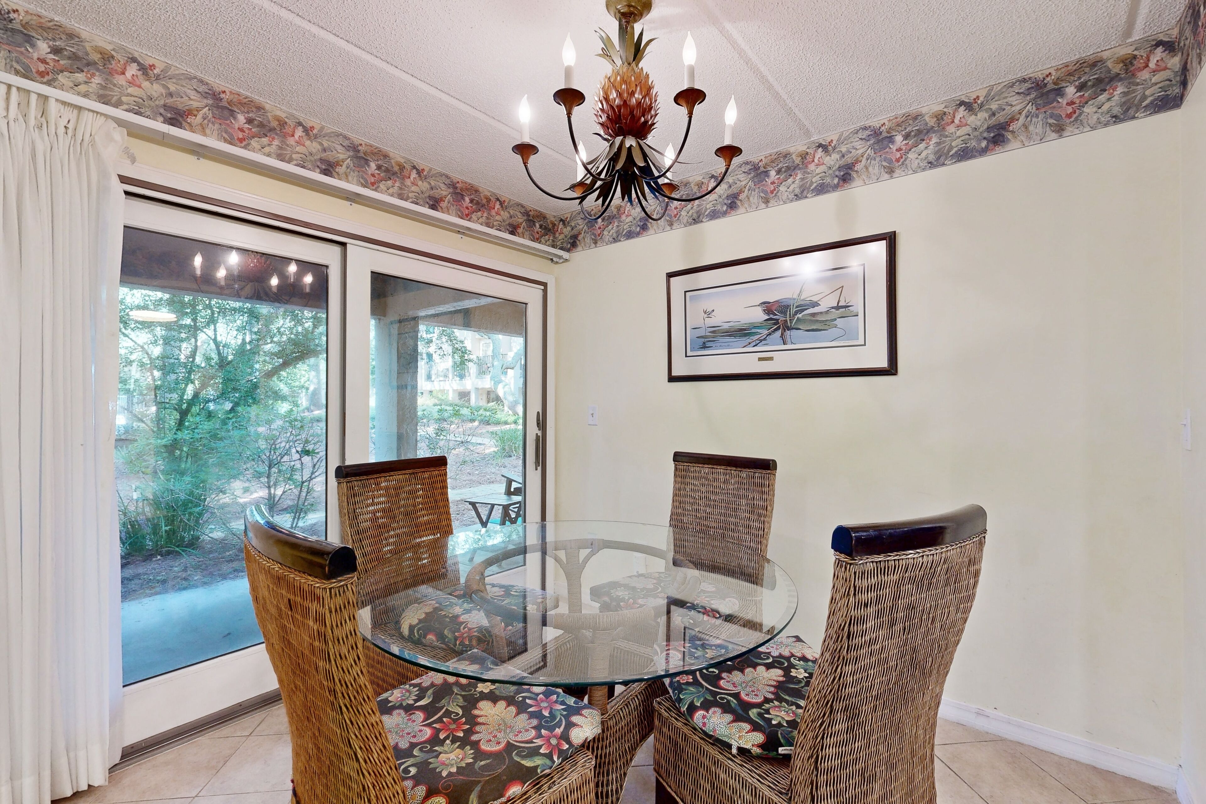 A dining area with a glass table surrounded by wicker chairs with floral cushions, a chandelier, and a framed picture on the wall.