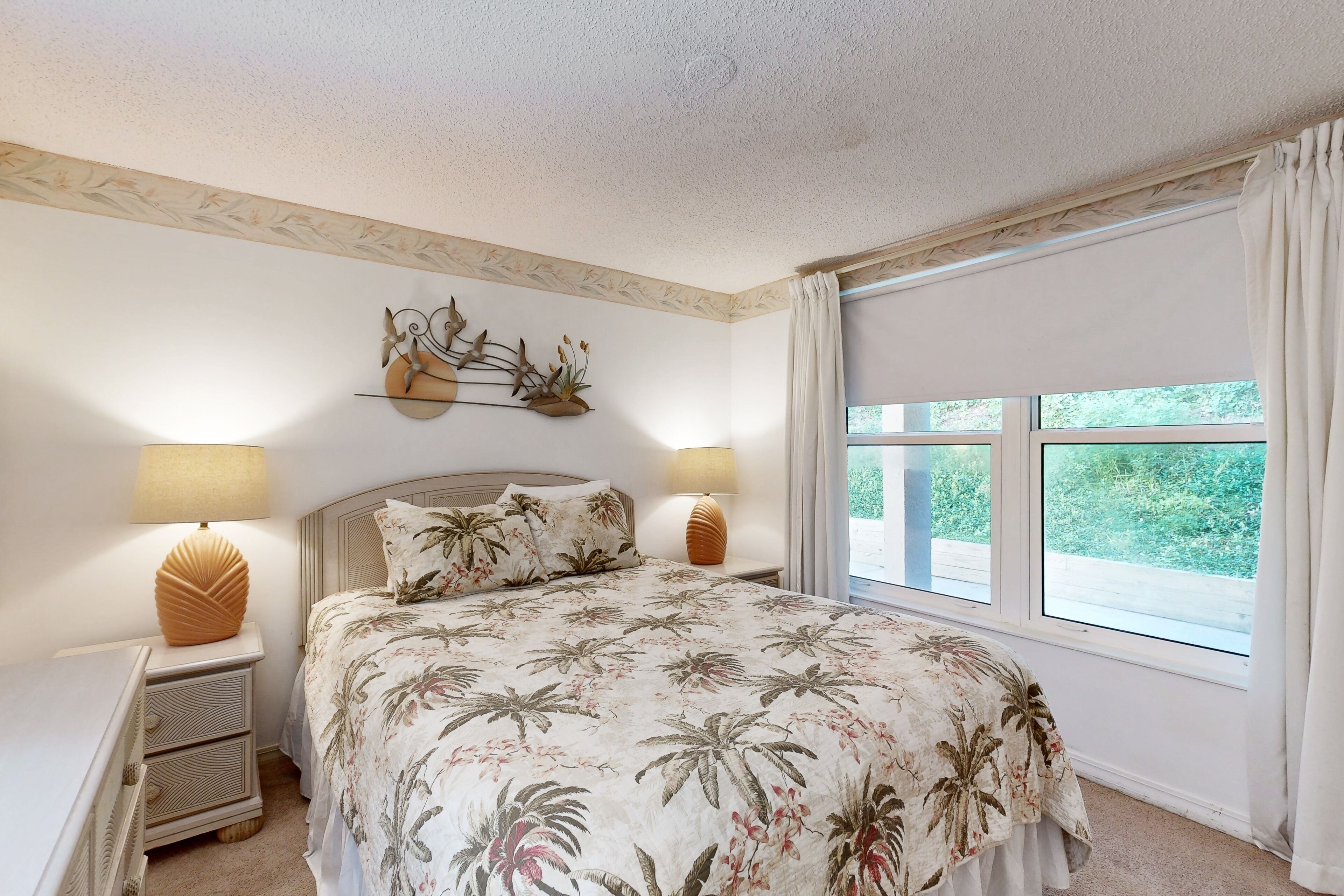 A bedroom with a bed, two bedside lamps, a nightstand, a decorative wall piece, and a window with curtains.
