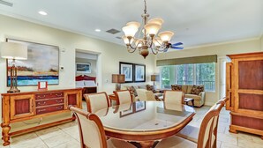 A spacious dining area with a large wooden table and chairs, a chandelier, and a view of the outdoors through large windows. - Dog-friendly Home in the Tennis Villas W/free Wifi, Shared Pool, Central AC (Fernandina Beach)