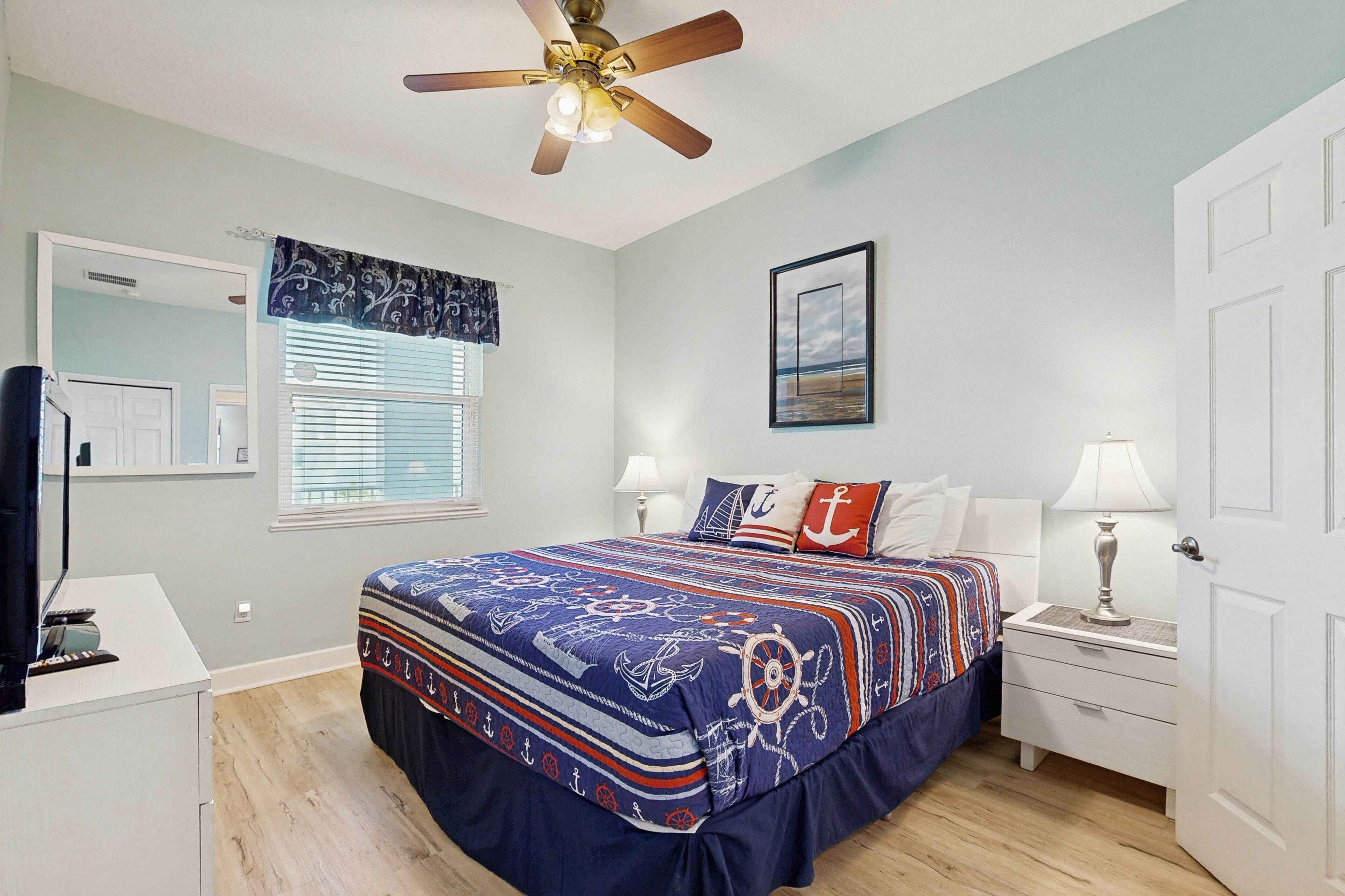 A bedroom with a bed, nightstands, a desk, a ceiling fan, a window with blinds, and a door.