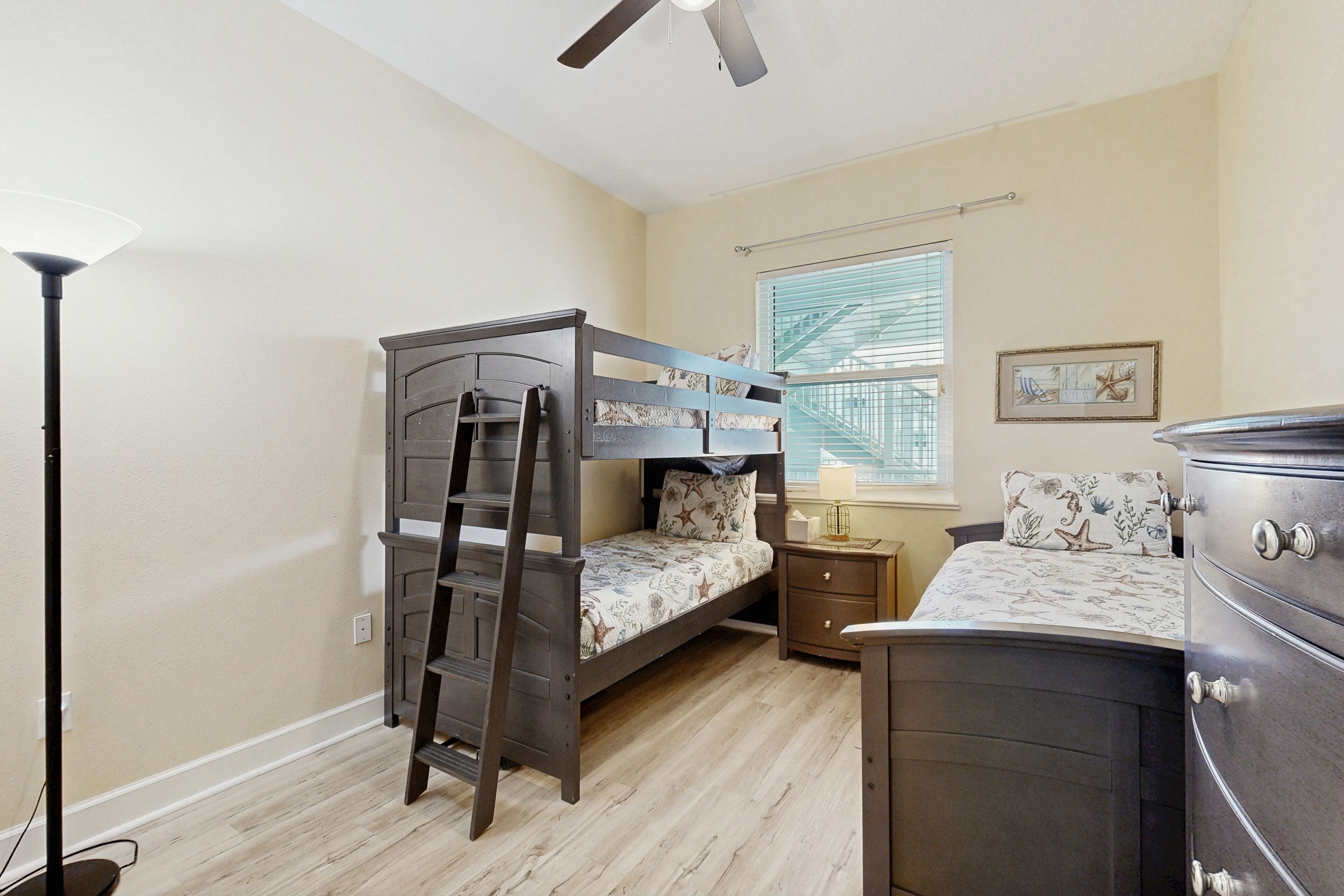 A bedroom with a bunk bed, a dresser, a nightstand, a lamp, a window with blinds, and a ceiling fan.
