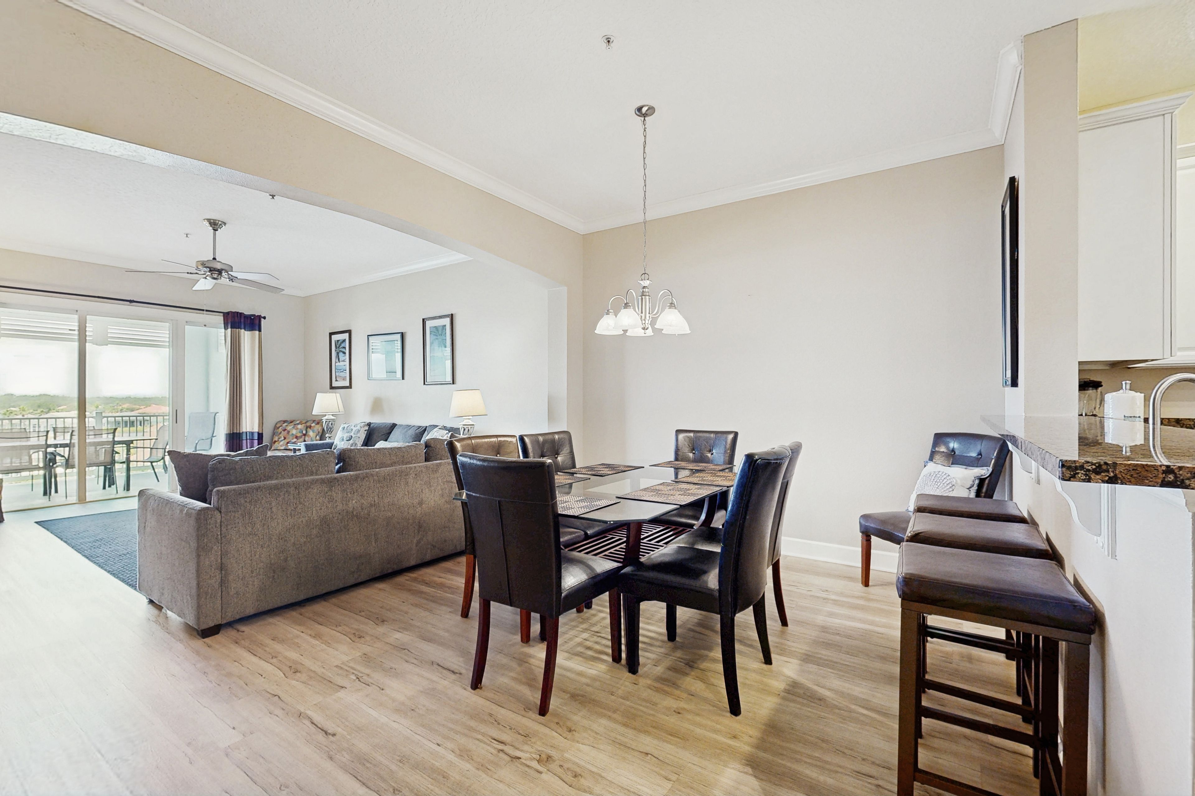 A spacious living room with a large sofa, dining table, and chairs. There is a balcony with a view, and a kitchen area with a countertop and sink.