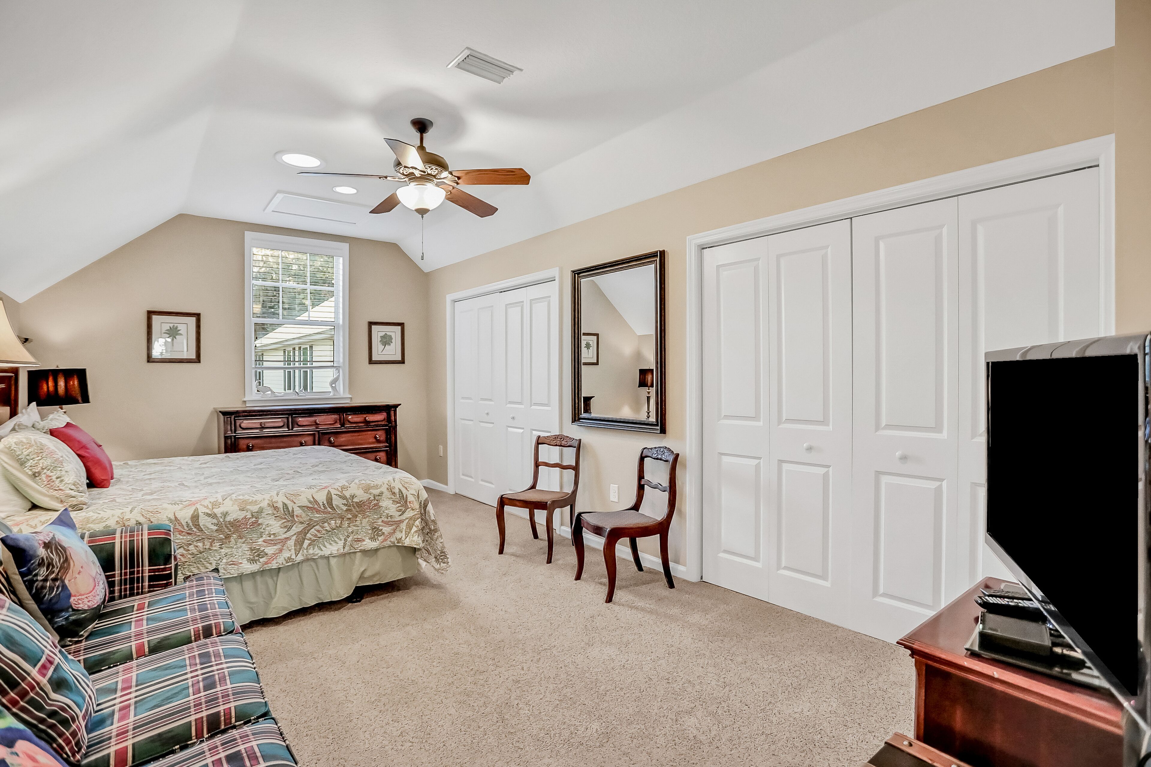 A bedroom with a bed, two chairs, a TV, and a ceiling fan.