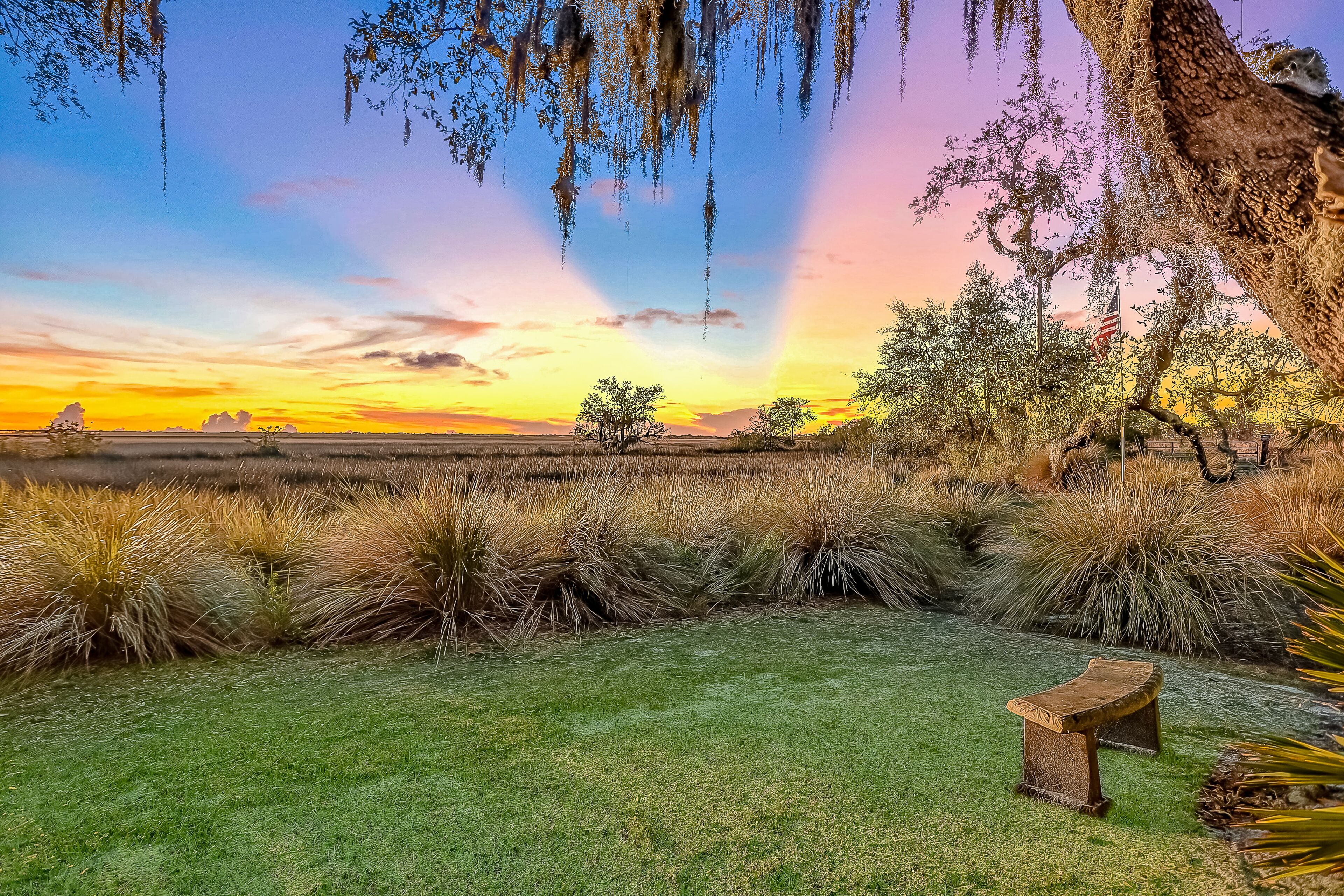 A grassy field with a wooden bench, trees, and an American flag.