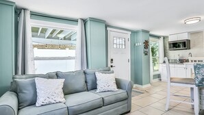 Una sala de estar con un sofá gris, una puerta blanca y una zona de cocina con microondas y fregadero.