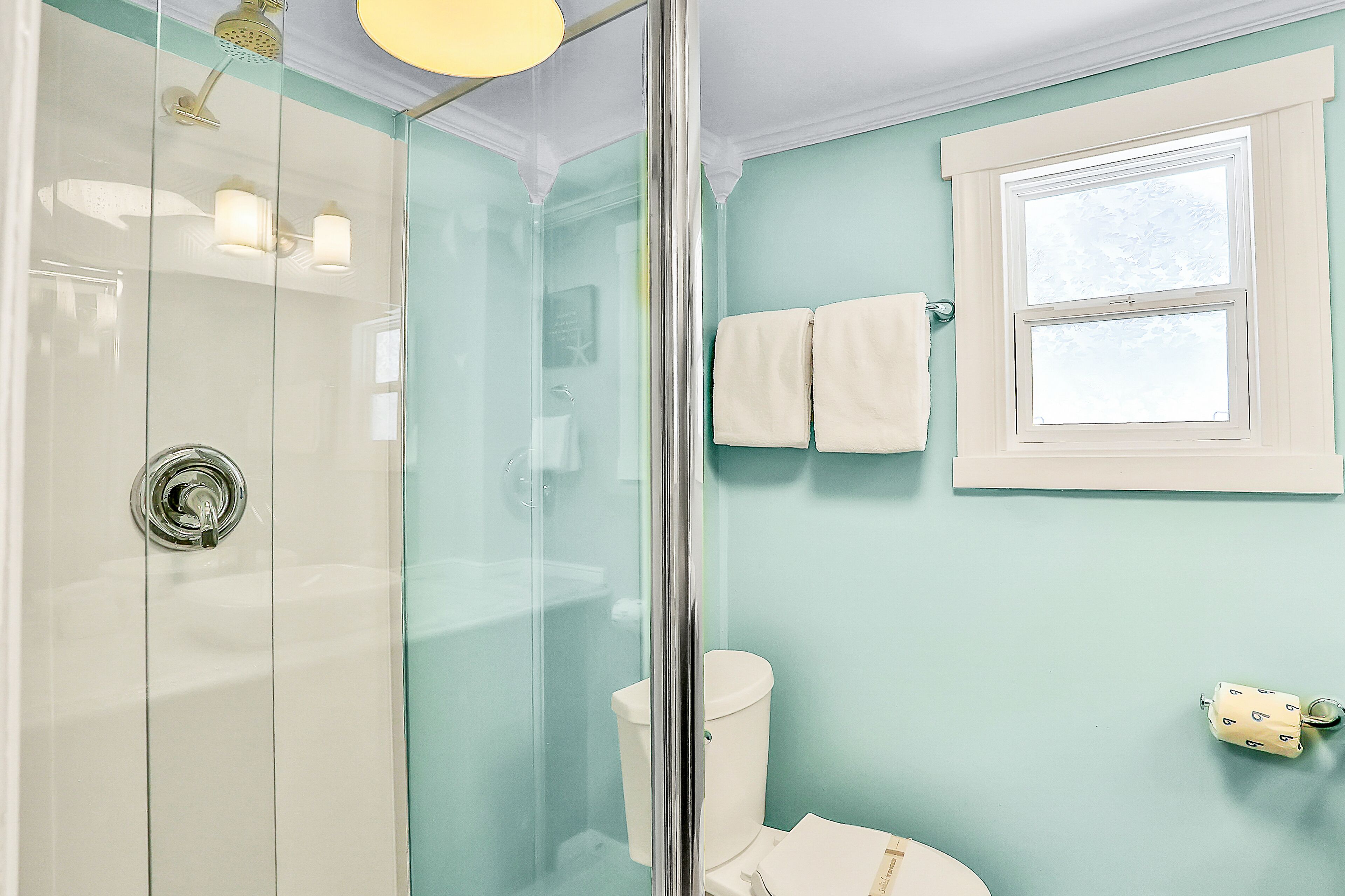 Ein Badezimmer mit Duschkabine aus Glas, Toilette und einem Fenster, an dessen Wand Handtücher hängen.
