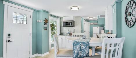 A compact kitchen with white cabinets, a microwave, and a refrigerator. There is a dining area with a table and chairs, and a clock on the wall.