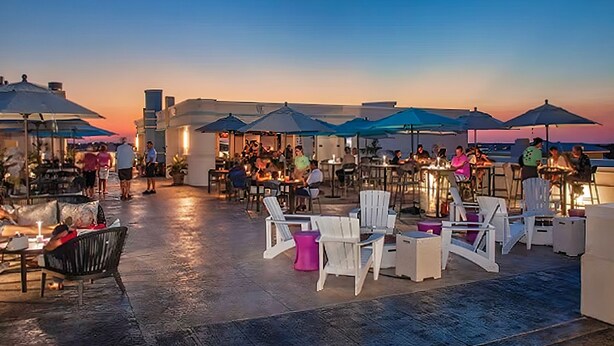 Un área de comedor al aire libre con mesas, sillas, sombrillas y una puesta de sol al fondo.