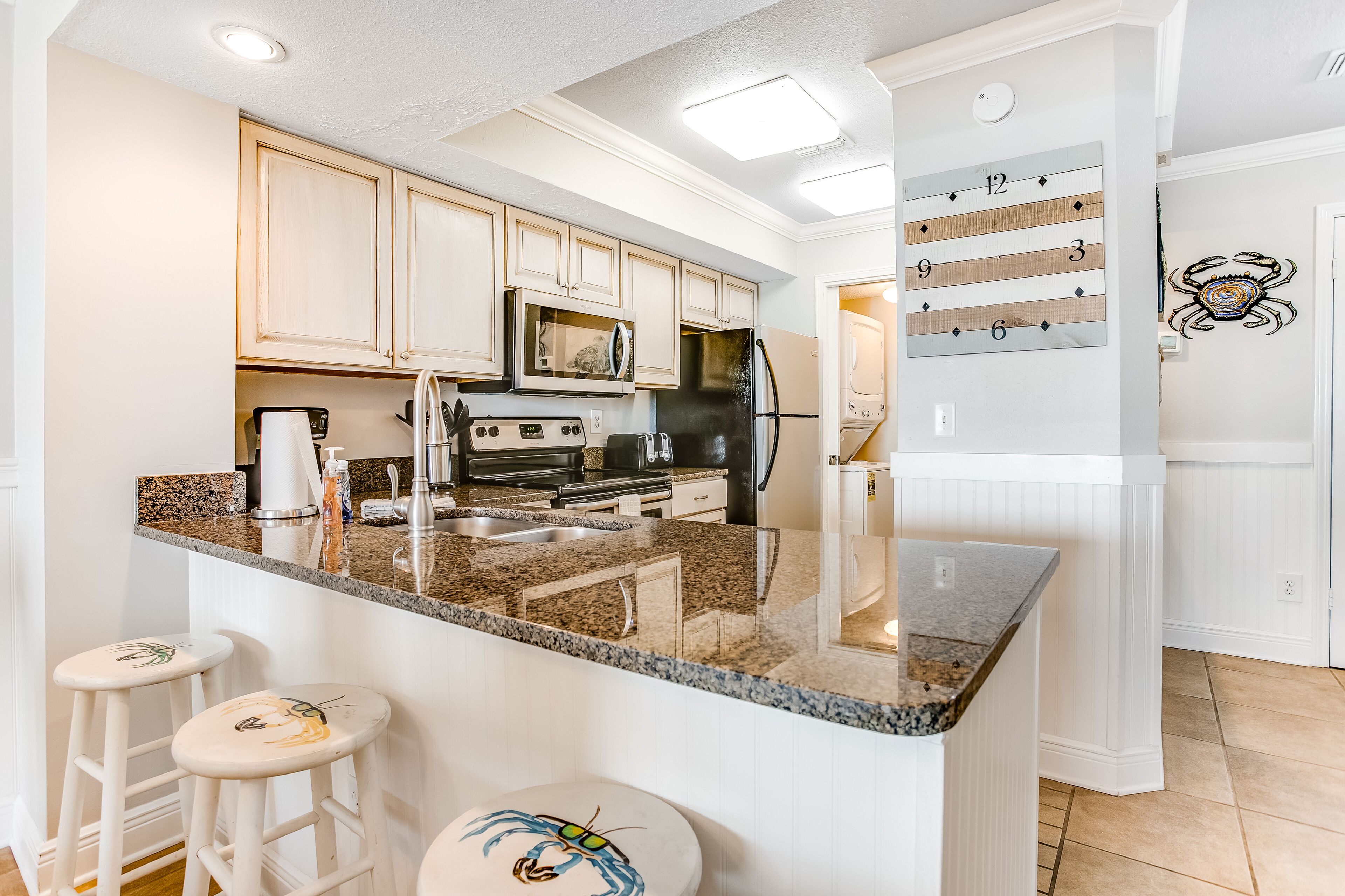 A kitchen with white cabinets, a granite countertop, a refrigerator, a microwave, and a sink. There are two stools with crab designs.