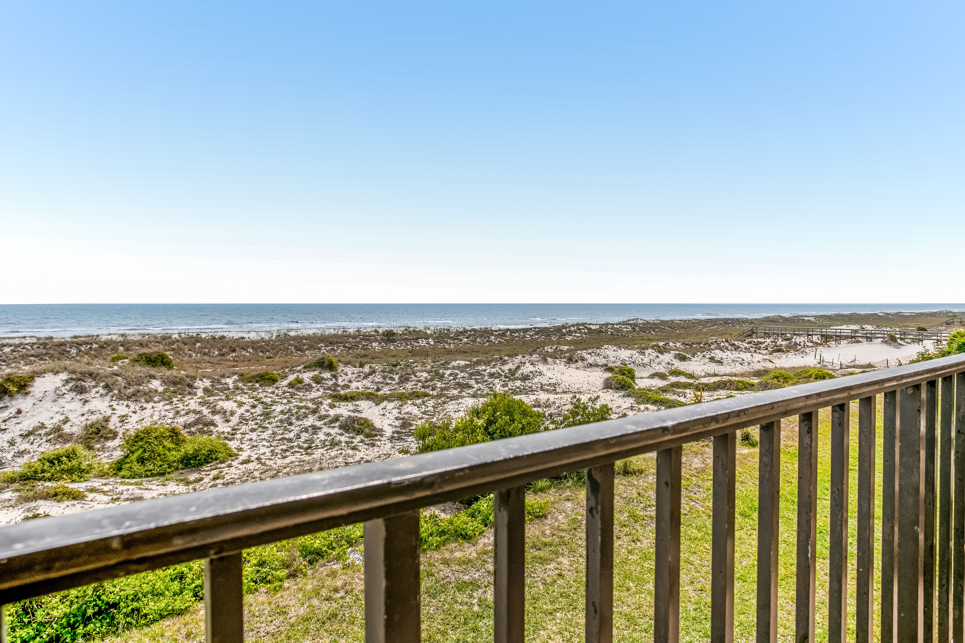 Blick auf einen Strand vom Balkon mit Holzwertung, feiner Sand und vereinzelte Vegetation.