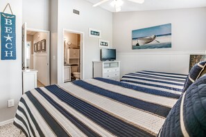 Quarto com colcha listrada, televisão sobre suporte e quadro com tema de praia na parede.