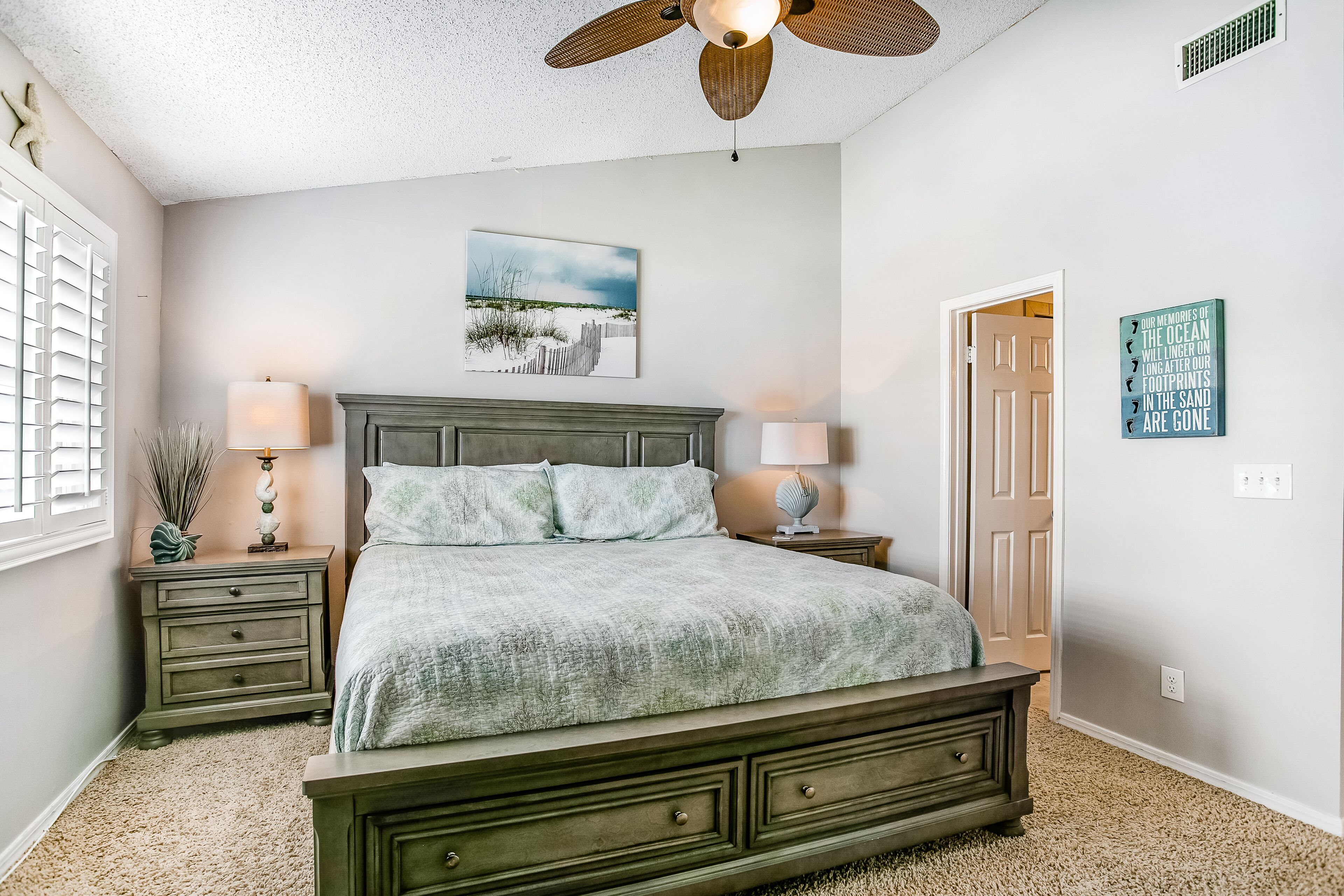 A bedroom with a bed, nightstands, a ceiling fan, a window with blinds, a door, and a wall with a picture and a sign.
