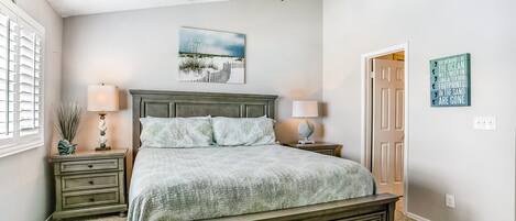 Une chambre à coucher comprenant un lit, des tables de chevet, un ventilateur de plafond, une fenêtre avec des stores, une porte et un mur orné d’un tableau et d’une affiche.