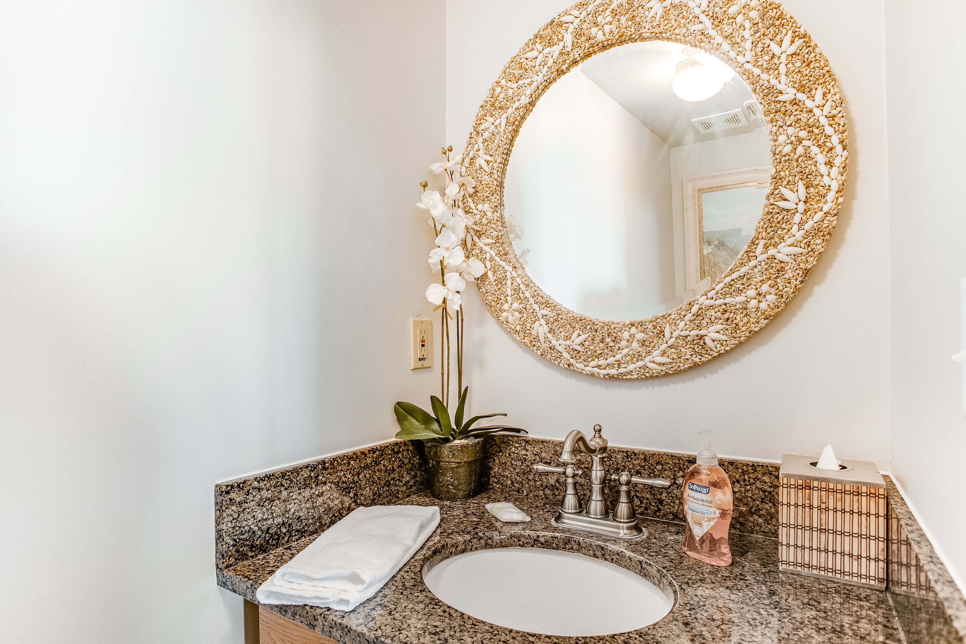 Un baño con encimera de granito, un lavabo blanco, un espejo con marco decorativo, una orquídea en maceta y una botella de jabón líquido.