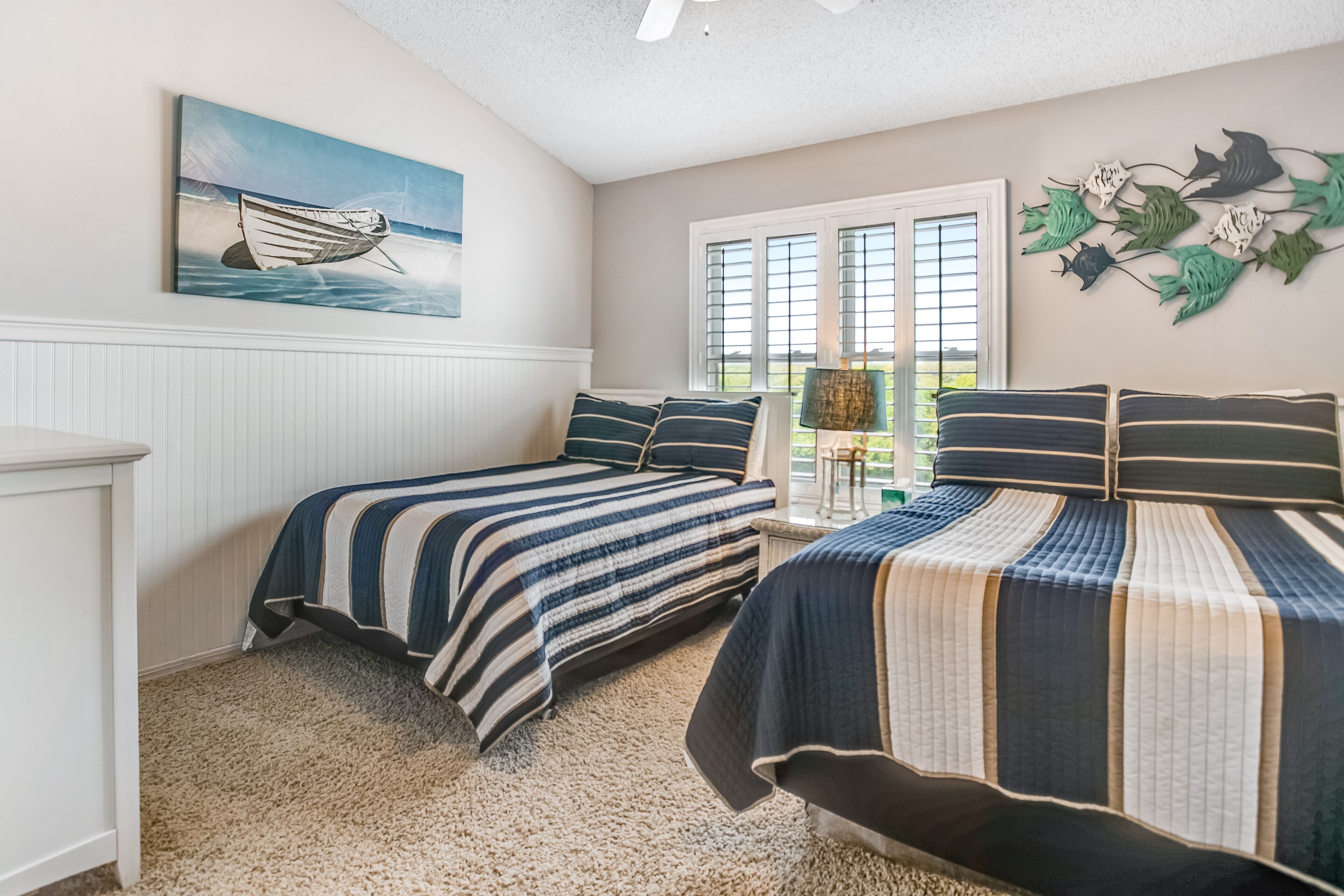 A bedroom with two beds, a window with blinds, a painting of a boat, and a wall decoration of fish.