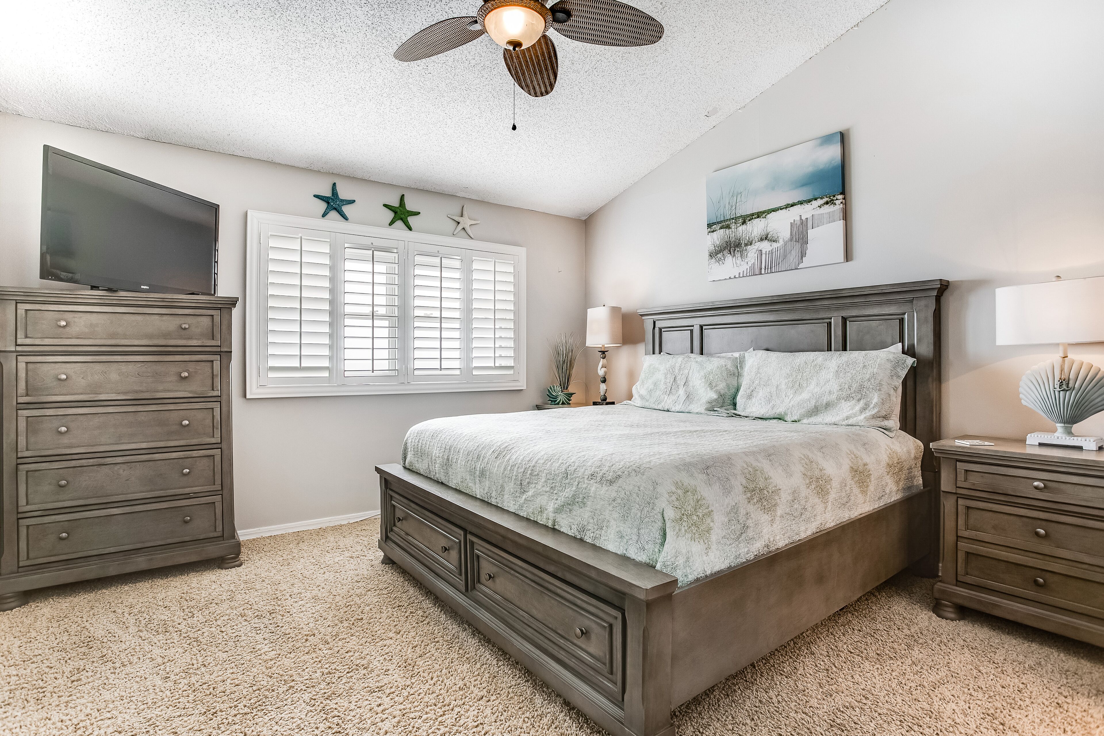 A bedroom with a bed, dresser, nightstand, ceiling fan, window with shutters, and wall decorations.