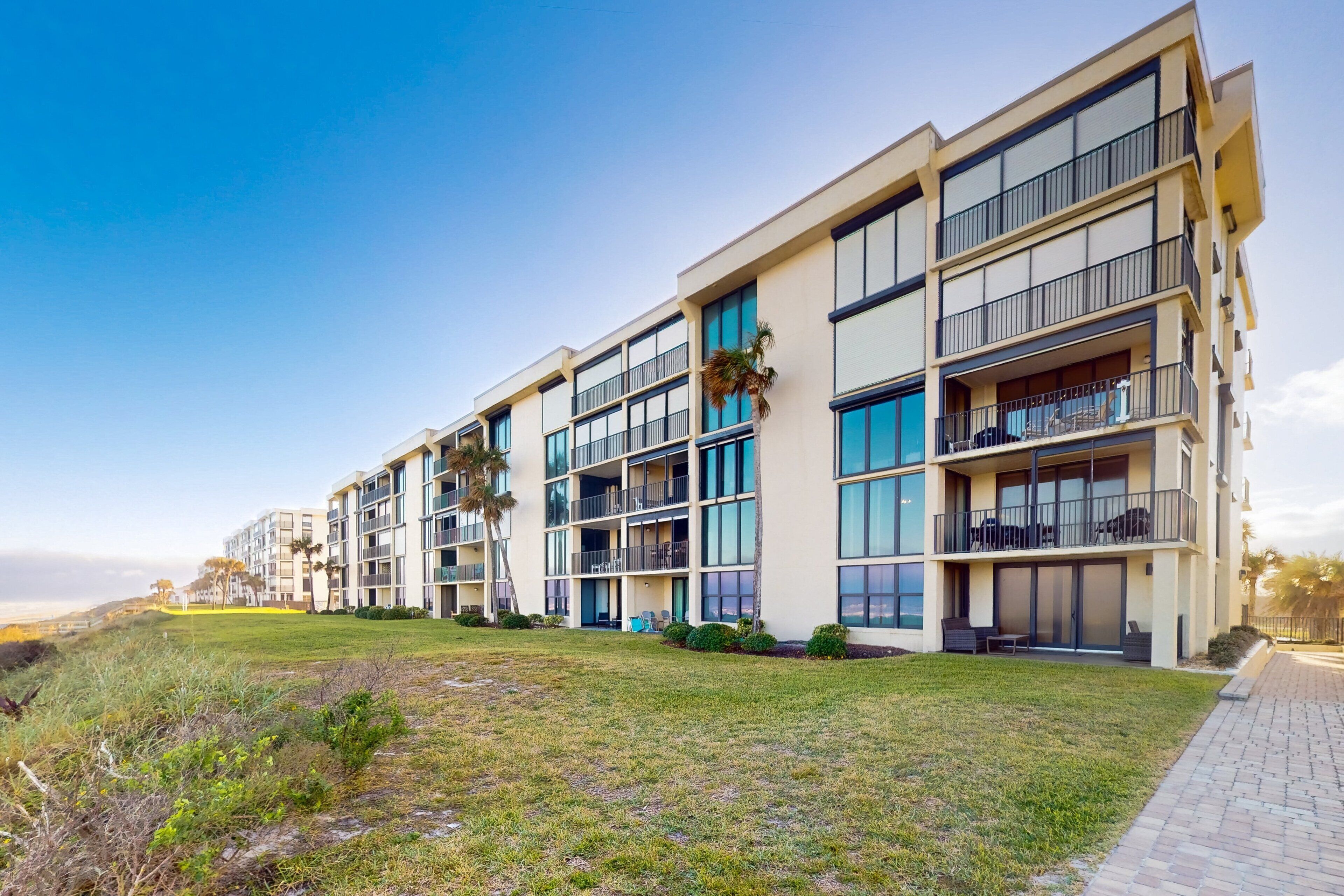 A multi-story hotel with balconies, large windows, and a grassy area in front.