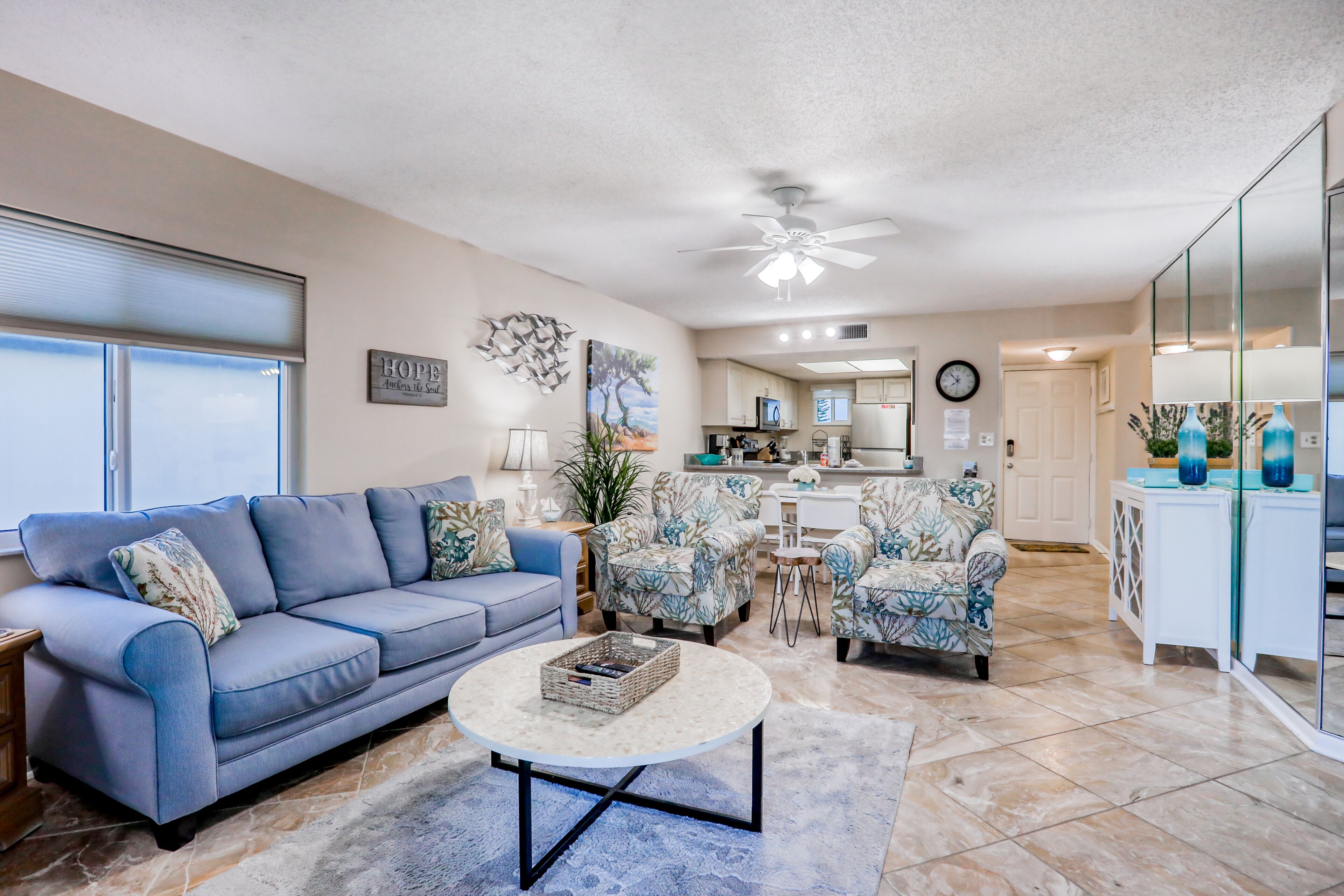 A living room with a blue sofa, a round coffee table, and floral-patterned armchairs. A kitchen is visible in the background with a dining area.