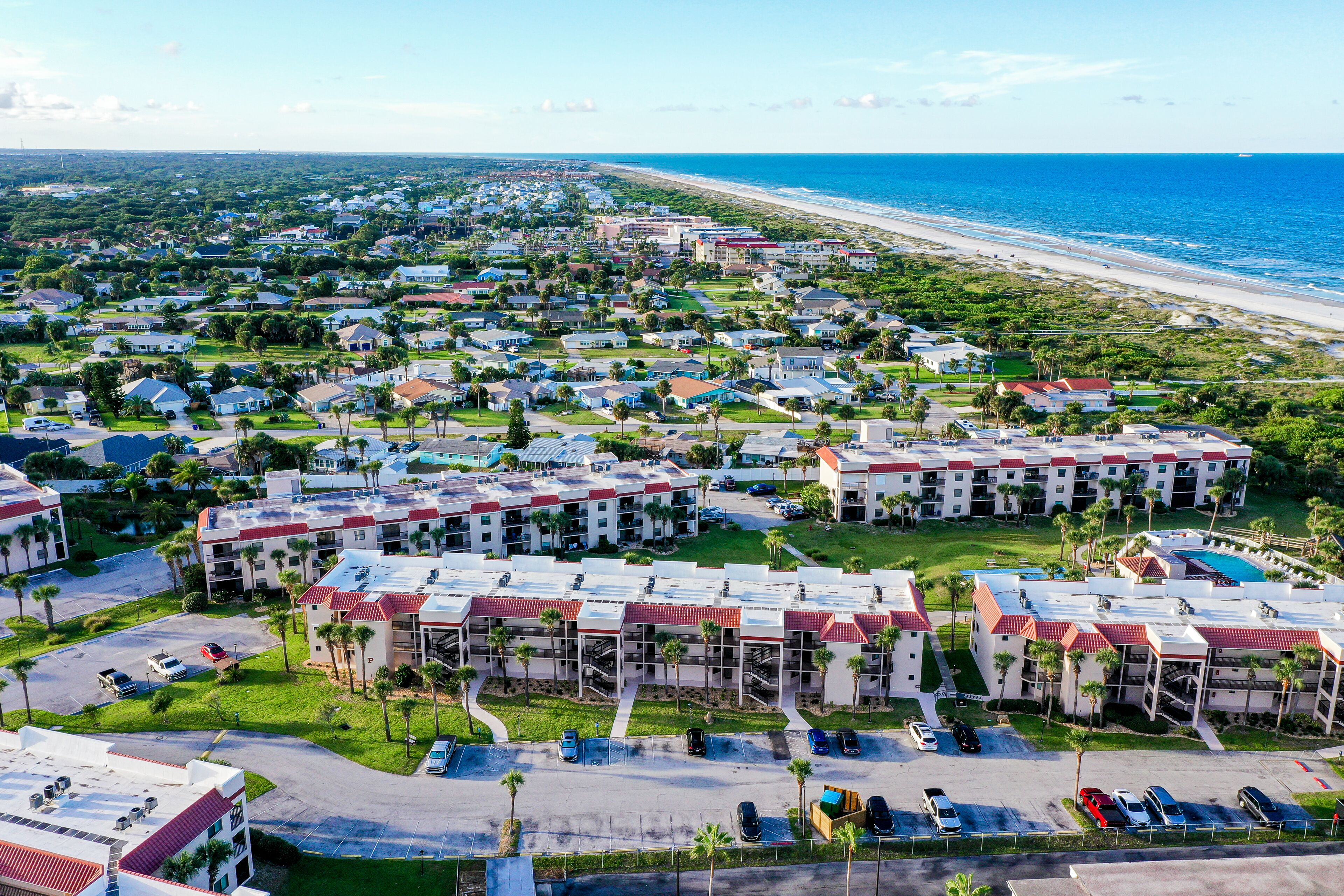 Vista aérea de una zona residencial costera con un hotel frente a la playa, que cuenta con varias construcciones, estacionamiento y abundante vegetación.