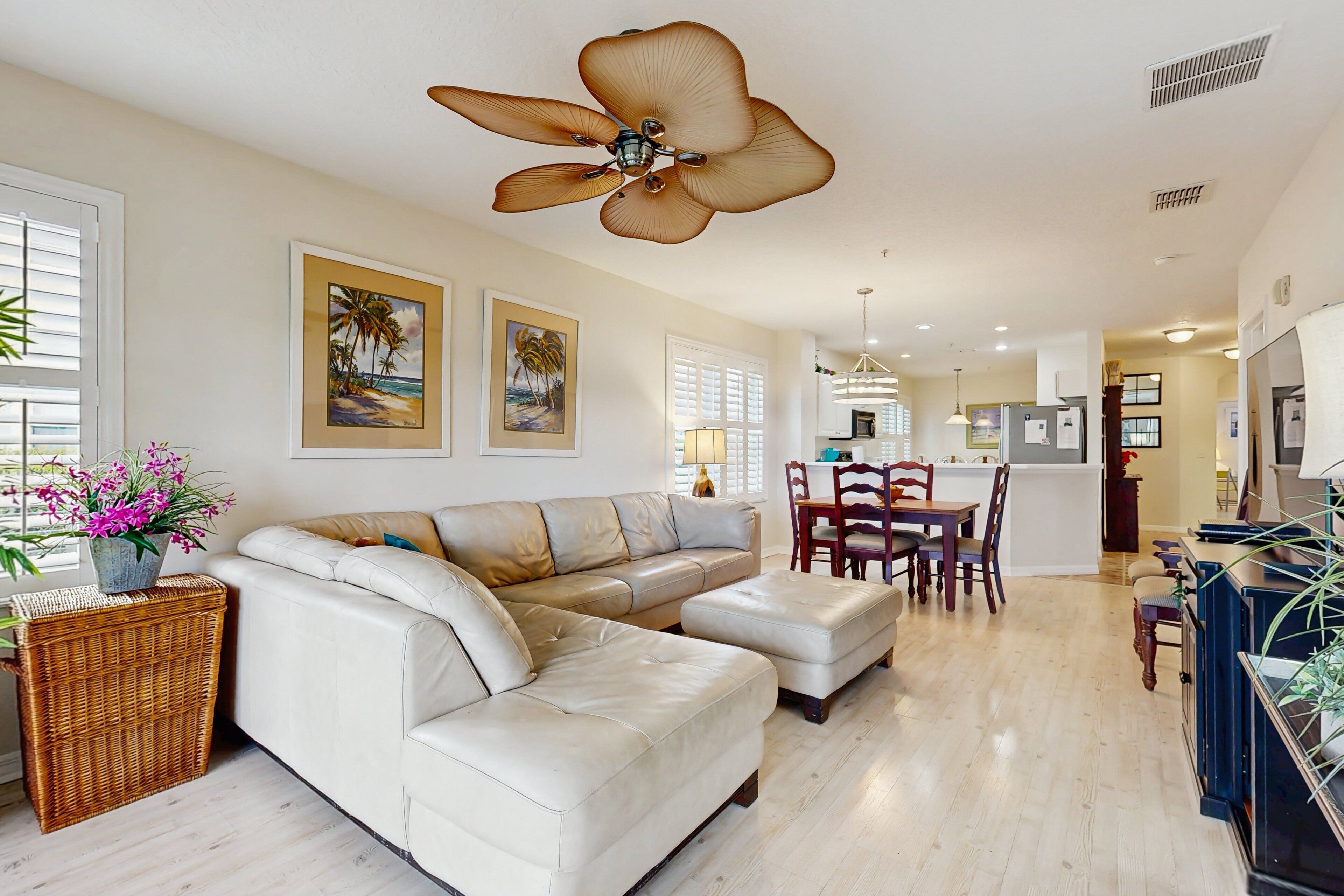 A spacious living room with a white leather sectional sofa, a dining area with a wooden table and chairs, and a ceiling fan with a large flower design.