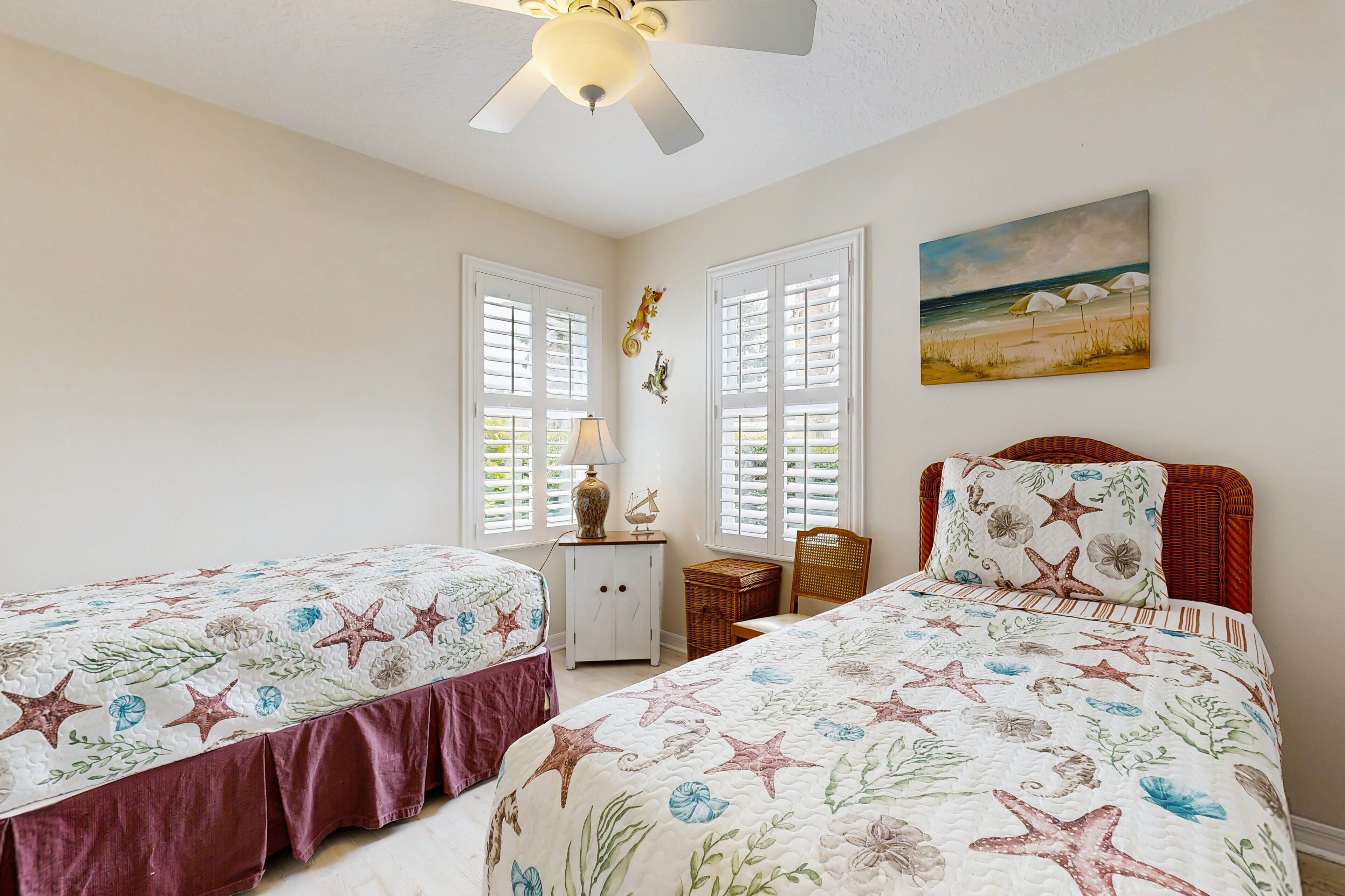 A bedroom with two beds, a ceiling fan, a painting of a beach scene, and a window with shutters.