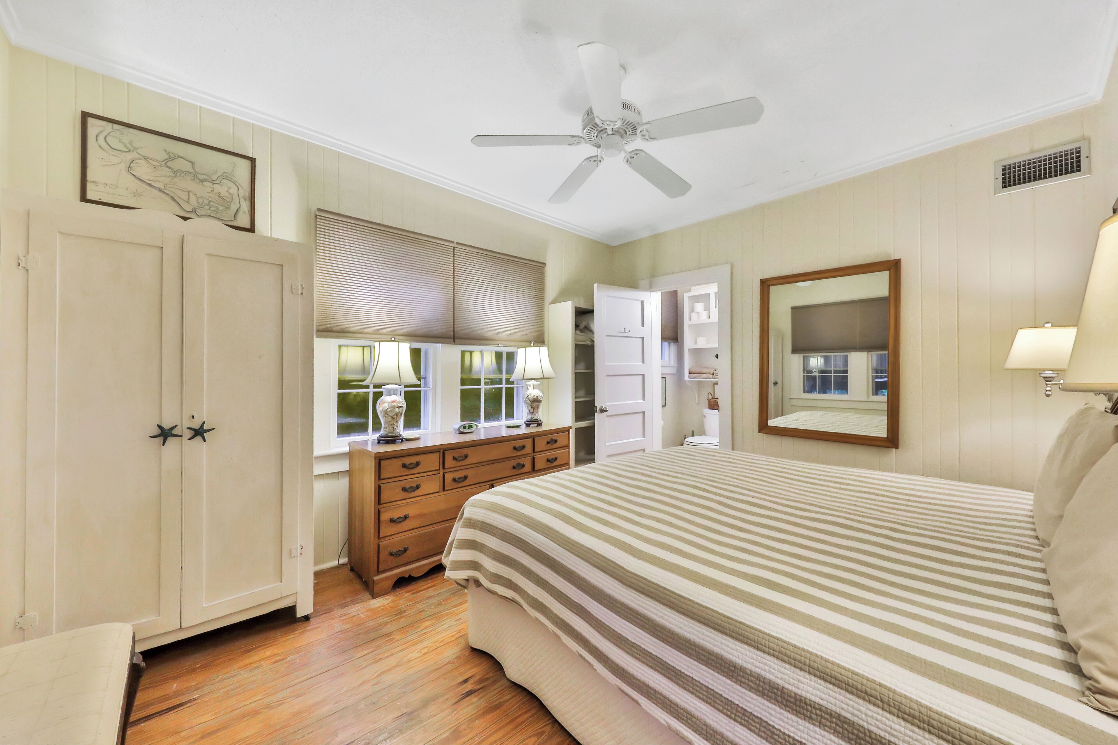 A bedroom with a bed, a dresser, a vanity, a ceiling fan, and a window with blinds.
