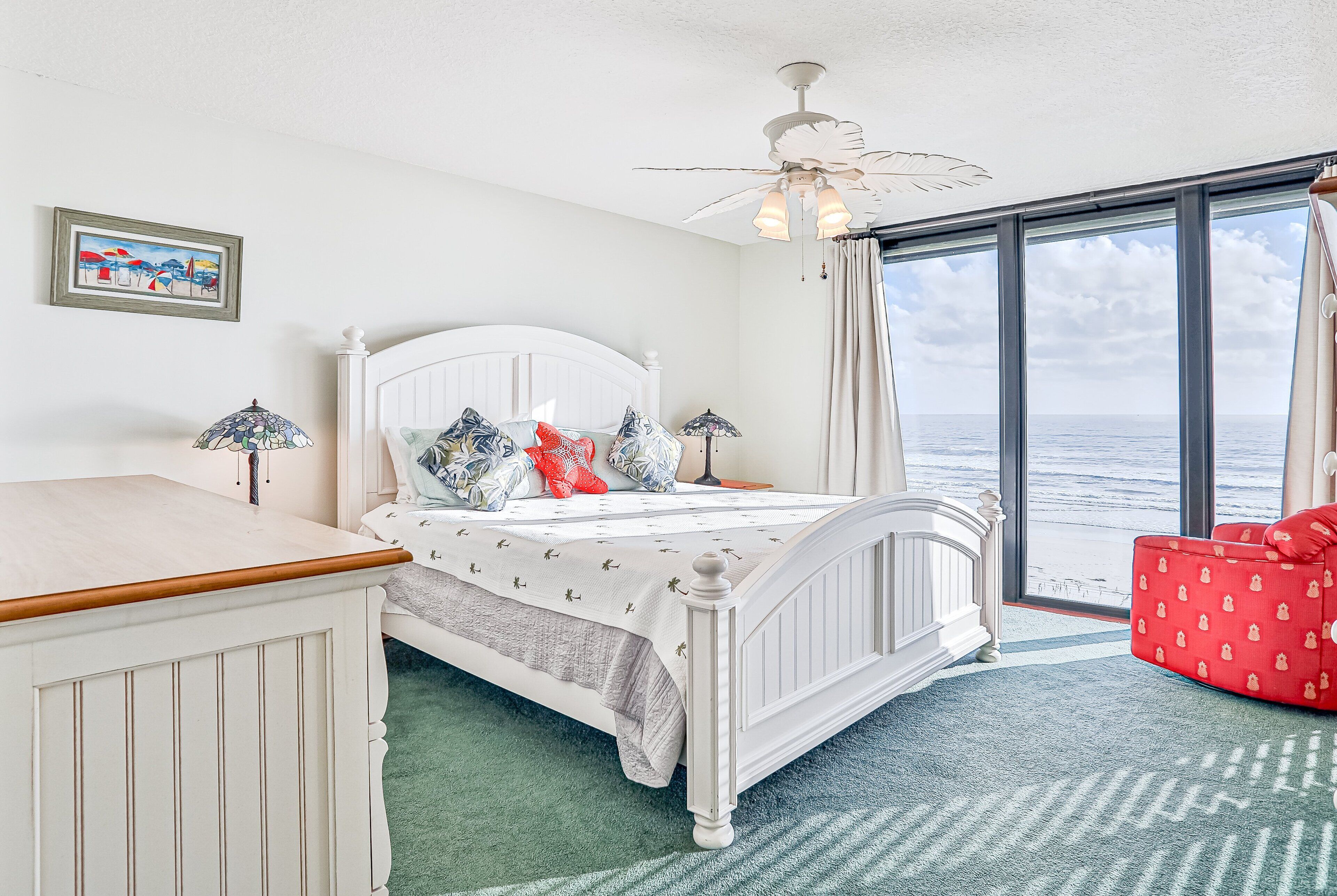 A bedroom with a bed, a desk, a chair, a ceiling fan, and a large window with a view of the sea.