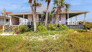 Een huis aan het strand met een overdekte veranda, palmbomen en een zandrijk gazon.