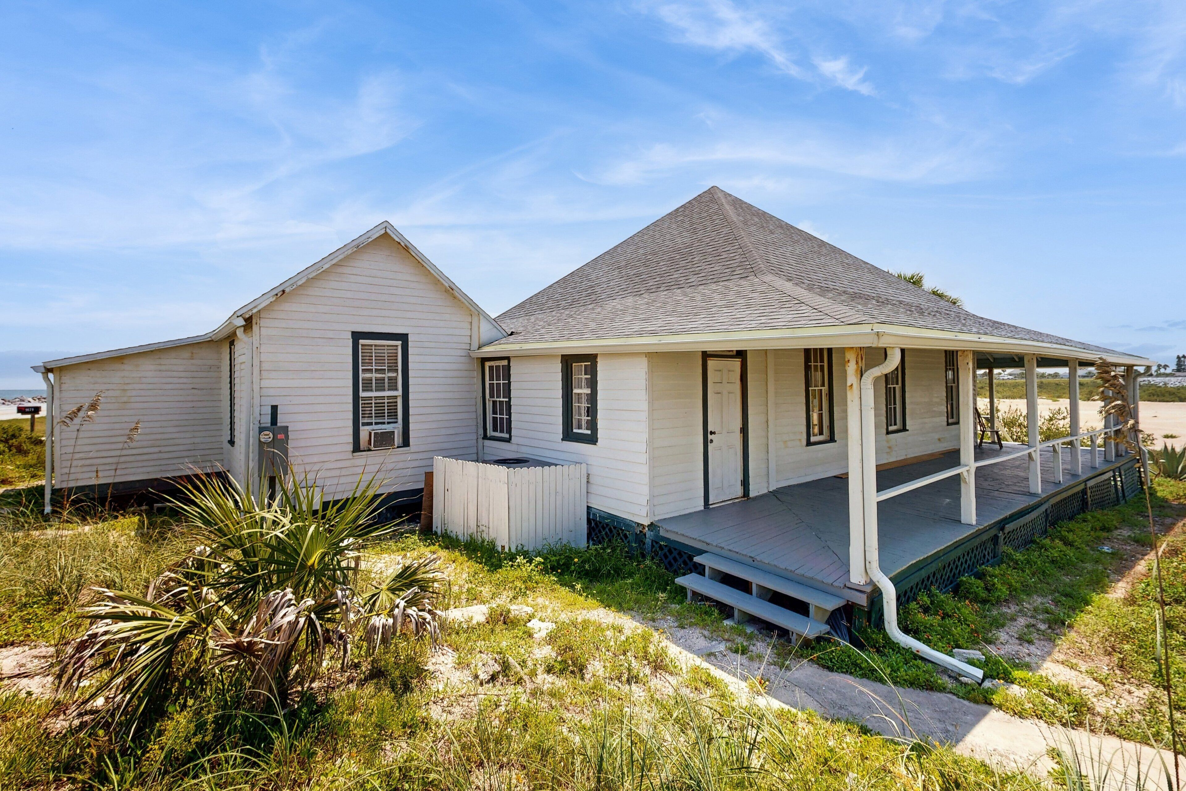 A two-story white building with a porch and a small deck, surrounded by grass and vegetation.
