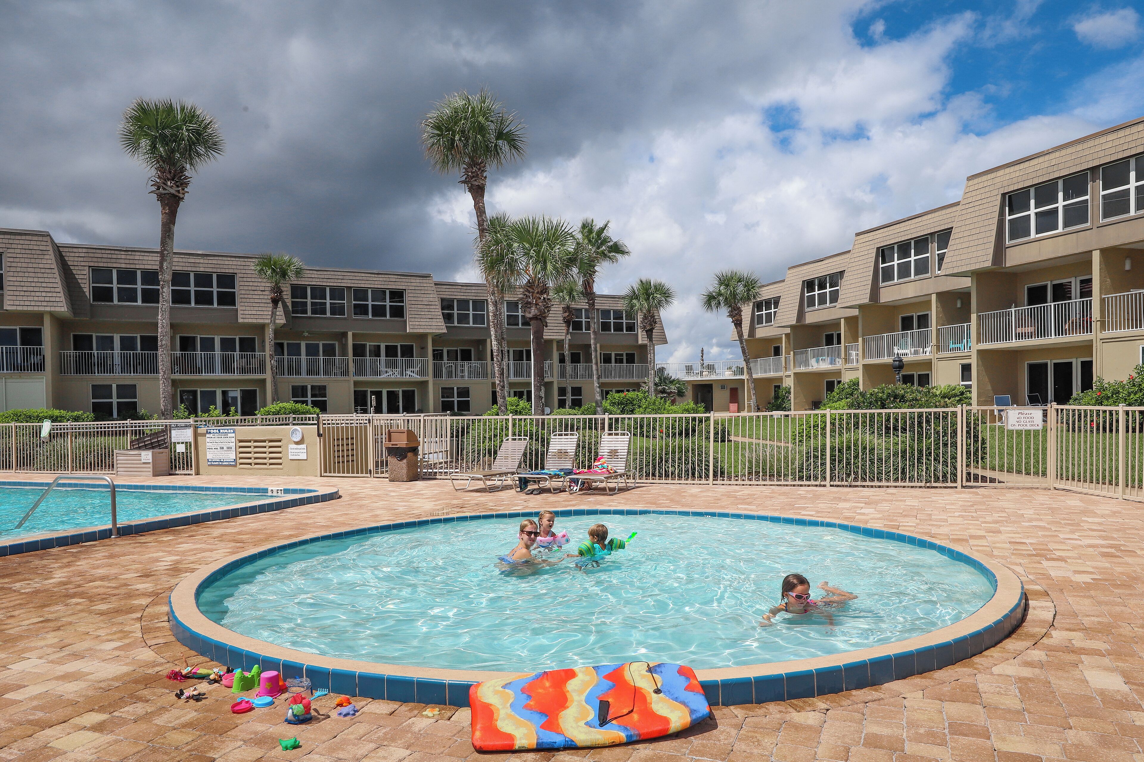 A swimming pool area with multiple pools, palm trees, and a building with balconies.